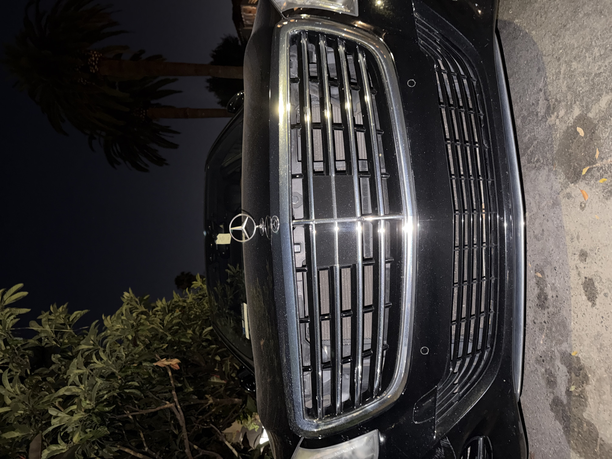 An extreme low-angle, close-up, full-shot photograph captures the front grille and hood of a black Mercedes-Benz sedan at night in an outdoor setting. The car is parked on a textured, light-colored surface that appears to be asphalt or concrete, with some dark wet patches and scattered fallen leaves. The Mercedes-Benz logo, a silver three-pointed star within a circle, is prominently displayed on the hood, above the large, chrome-accented grille. The grille itself is composed of horizontal slats with a mesh backing, reflecting light from an unseen source. The car's headlights are visible on either side, and a sensor is embedded in the lower part of the bumper. Behind the car, a dark sky is visible, with the silhouettes of lush green trees and palm trees, hinting at a warm climate. The lighting suggests artificial illumination or streetlights, casting subtle reflections on the car's surface and creating deep shadows.
