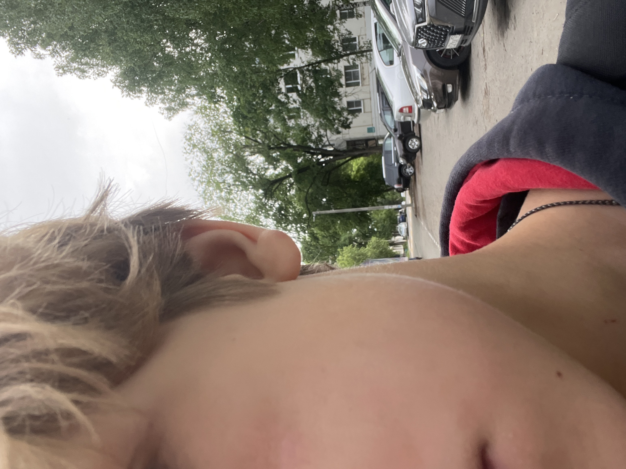 The image shows a close-up perspective of a person's face and the surrounding environment. Part of their face is in focus, including the ear, hair, and partial view of the eye and nose. The hair appears to be blonde.
In the background, several cars are parked along a street. Beyond the cars, there are trees, a building and an overcast sky.
It is not possible to confirm the location in the city of Chaikovsky, Russia, from the image alone.