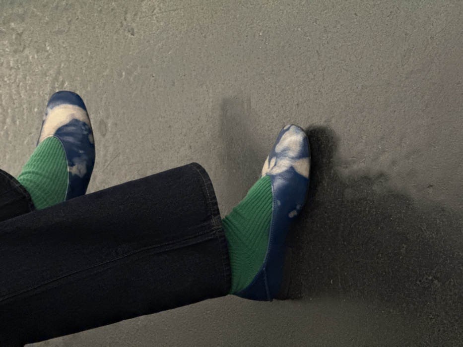 The image displays the lower extremities of an unidentified individual. The subject wears dark blue trousers, bright green ribbed socks, and flat shoes with a distinctive blue and white pattern resembling tie-dye or mottled clouds on the toe box and upper. The individual's left foot is positioned slightly higher and further back than the right. Both feet rest on a dark, smooth, and moderately reflective floor surface, potentially polished concrete or stone. A plain, dark, vertical surface serves as the background. No specific location within Paris, France, or any other city, can be determined from the visual information provided.
