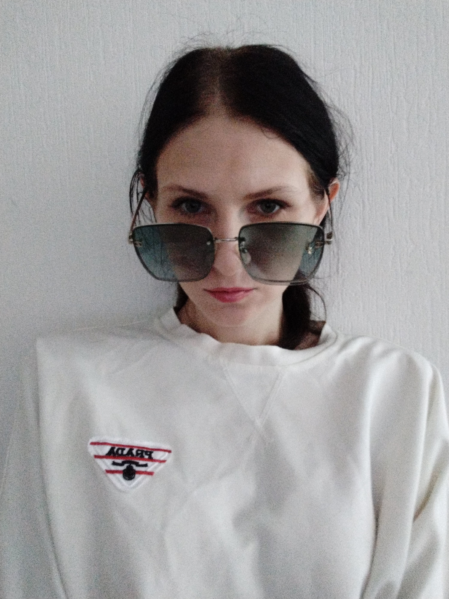A young woman with dark hair pulled back and wearing large, square, mirrored sunglasses stands against a plain white wall. She is looking directly at the camera, with her mouth slightly open and red lipstick visible. She is wearing a white crewneck sweatshirt with a triangular logo on the left chest that appears to be embroidered and features red and black stripes and lettering that reads "PRADA" upside down. The lighting is soft and diffuse, creating a somewhat grainy effect on the image. The background is entirely out of focus.