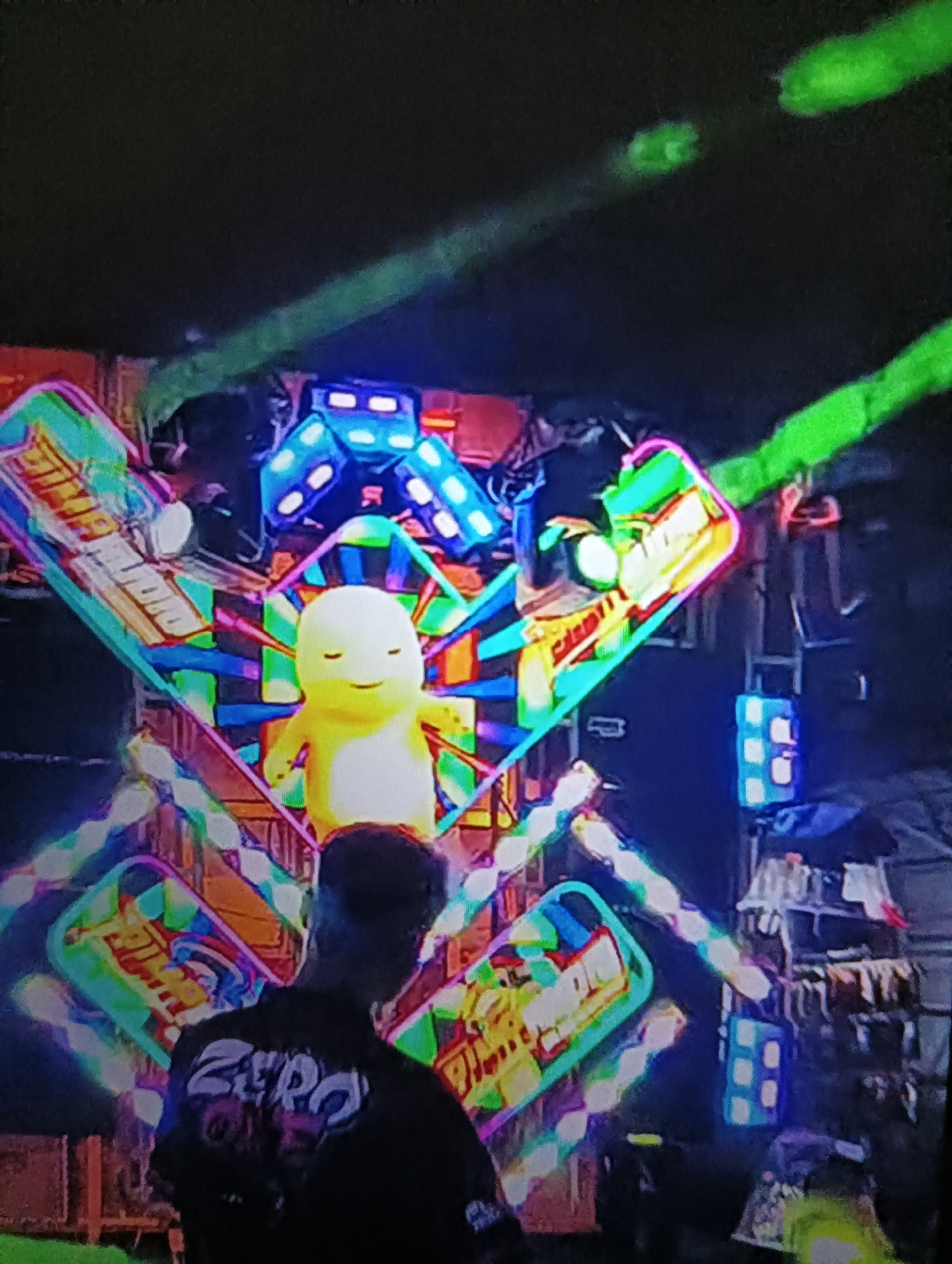 A nighttime outdoor scene features a person wearing a black t-shirt with a white graphic on the back, standing in front of a vibrant, neon-lit display. The display is composed of geometric shapes and frames, illuminated with bright green, blue, pink, and yellow lights. A cartoonish, yellow figure with closed eyes and a smile is prominently featured in the center of the display. The background is dark, with additional green light beams cutting across the frame from the top. The person is facing away from the camera, their head and shoulders visible. Some elements of the display are partially obscured by the person. The overall impression is of a festive or artistic installation, likely at an event or venue.