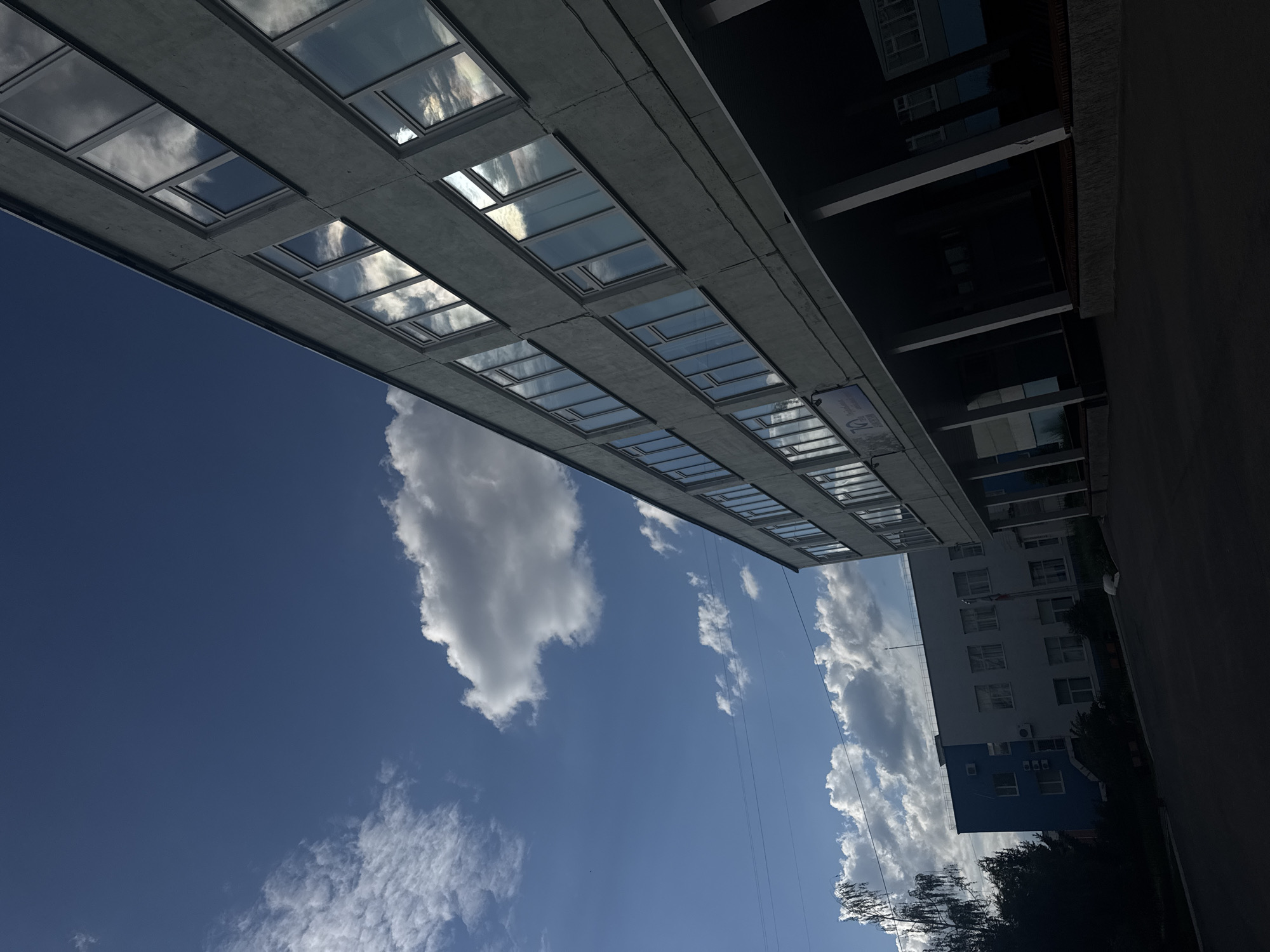 This media file depicts an exterior view of a modern building with a concrete facade and numerous rectangular windows. The windows reflect the sky, which is a bright blue with scattered white clouds. The image is taken from a low angle, looking up towards the building and the sky. To the right of the main building, another building is visible, partially obscured by shadows. This second building has a lighter facade and also features multiple windows. Trees with dark green foliage are present in the foreground on the right side of the image. The overall scene suggests a sunny day, possibly in the afternoon, given the bright sky and the quality of light. There is no discernible activity or people in the image. A sign with some text is visible on the lower right section of the concrete building, but the text is too small and blurry to be read. The location context is Chaikovsky, Russia.