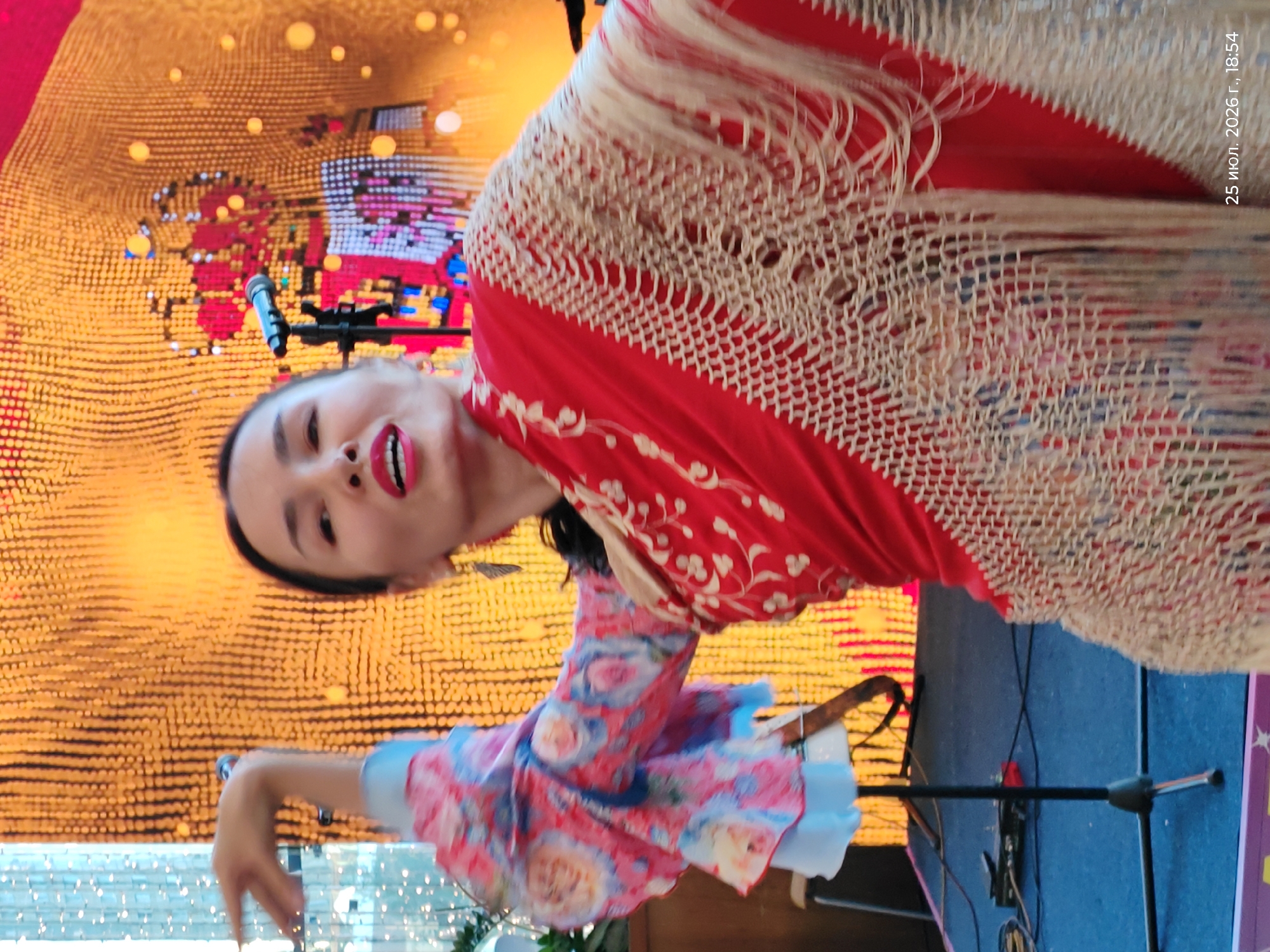 The image shows a woman dancing on a stage in Astana, Kazakhstan. The woman is wearing a red shawl with a fringed border and a colorful floral patterned blouse. She has her arms raised and is looking towards the audience. Behind her, a large screen displays a vibrant, abstract pattern of lights. A microphone stand is positioned to her right. The stage floor is blue, with some audio equipment visible. The overall scene suggests a lively performance or event.