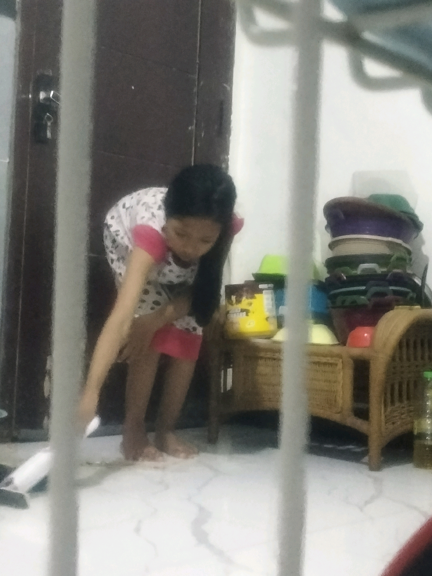 In Soko, Indonesia, a young girl with dark hair, wearing a white dress with black polka dots and pink trim, is bent over on a white tiled floor. She appears to be cleaning or picking something up. In the background, there is a brown door with a lock and a metal bar, and a wicker shelf unit holding stacked colorful plastic bowls and a yellow container.
