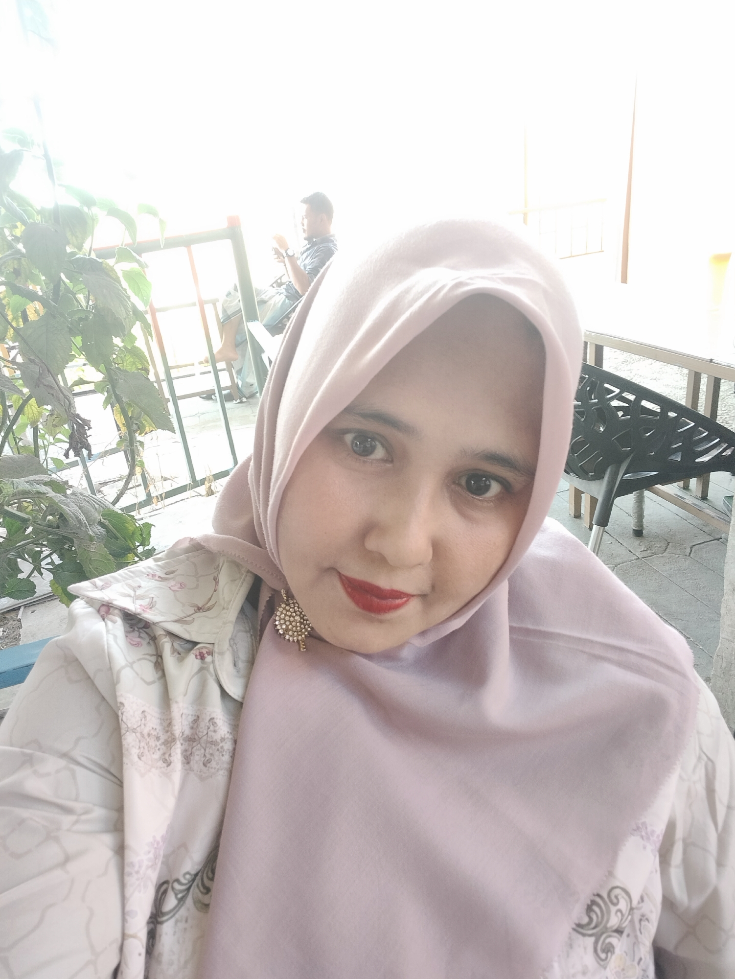 A person wearing a light purple hijab and white floral-patterned top is in a bright outdoor setting. The person has red lipstick and a sparkling earring. In the background, there is another person seated. A plant is visible to the left, and a black chair is to the right. The location appears to be outdoors, possibly a patio or open-air cafe in Matang, Indonesia.