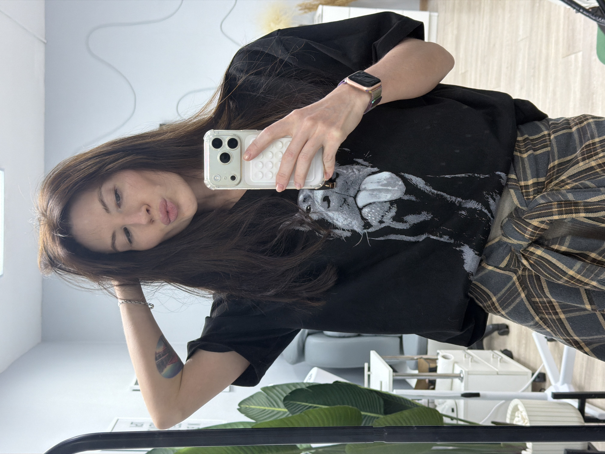 A woman is shown in a selfie, holding an iPhone with a clear case. She has dark brown hair and is wearing a black T-shirt with a grey wolf graphic and a grey and brown plaid flannel shirt. She has a smart watch on her left wrist and a bracelet on her right wrist. A green leafy plant is in the foreground, along with part of a black metal structure. The background shows a white wall, furniture, and a glimpse of what appears to be a medical or cosmetic treatment area. Based on available information, this photograph may have been taken in Perm, Russia.
