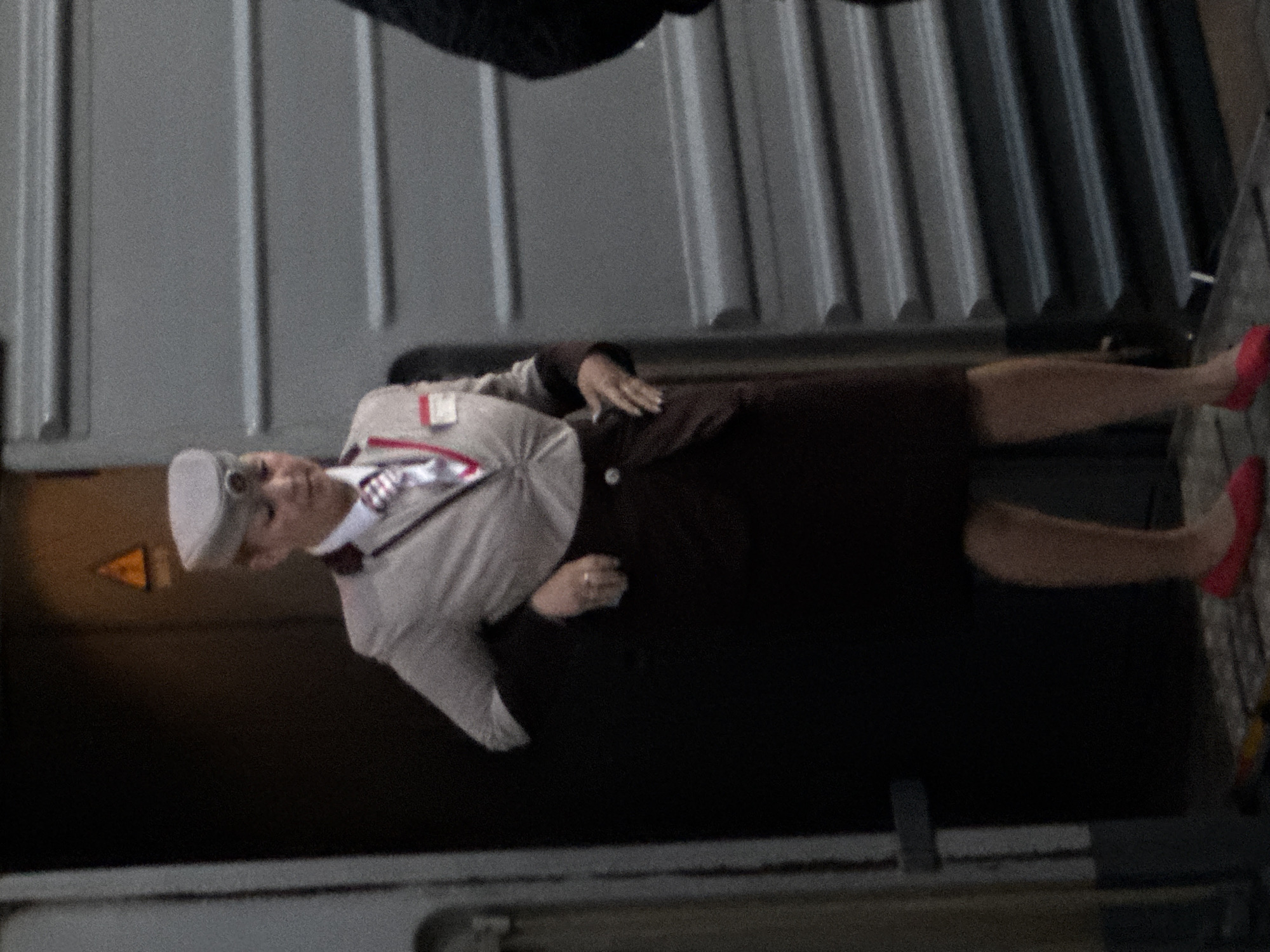 A woman wearing a gray uniform hat, a light-colored jacket with burgundy trim and a white collared shirt with a patterned tie, and a dark skirt stands next to an open train car door. She has a name tag on her left chest. Her right hand is resting on her hip, and her left hand is touching her skirt. She is wearing red flat shoes. The train car has gray ribbed siding. The background is dark and indistinct, with a yellow triangle warning sign visible in the upper left. The scene appears to be at a train station in Moscow, Russia.