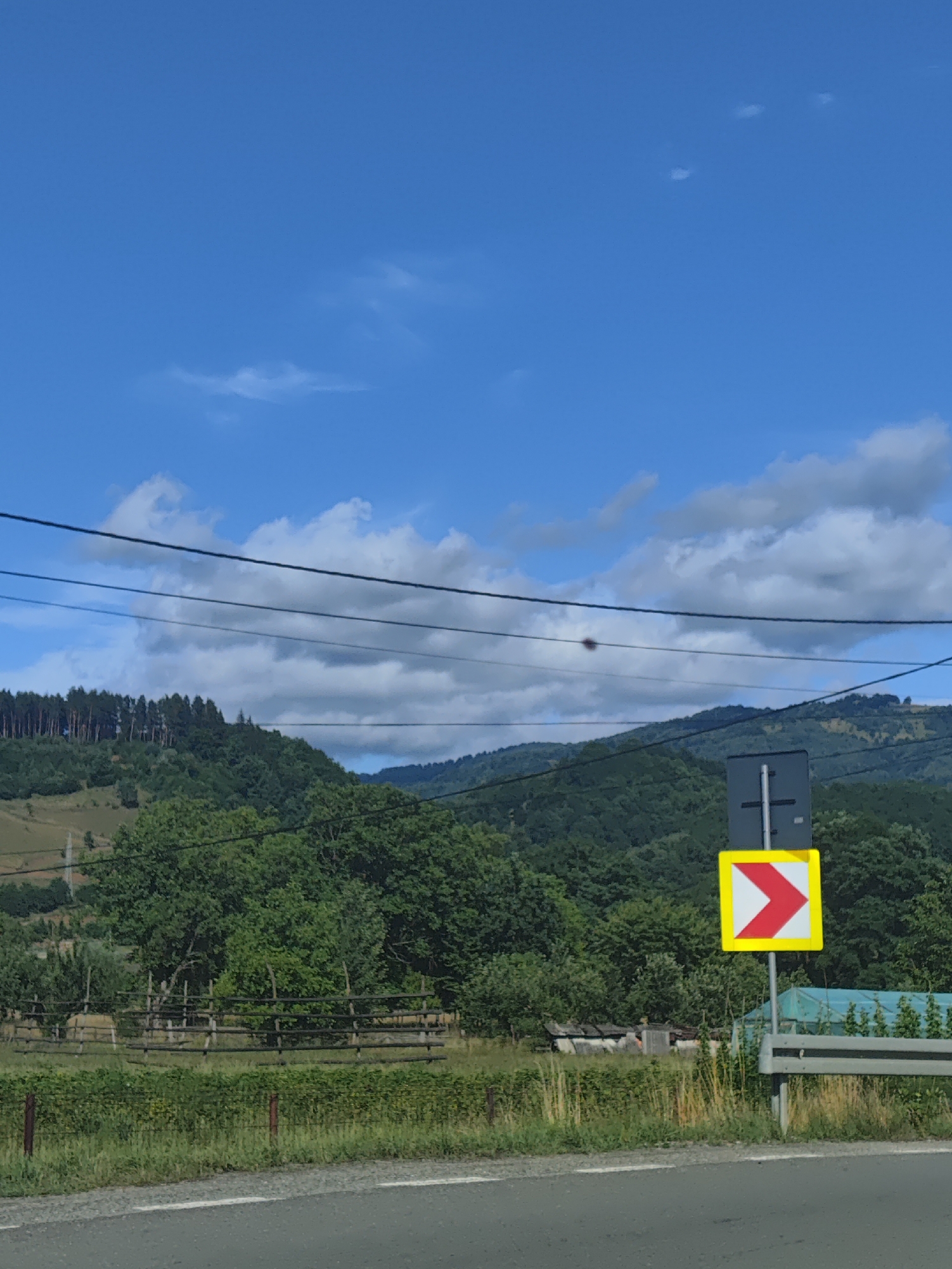 The image depicts a rural roadside scene, likely in the vicinity of Leordina, Romania, under a bright blue sky with scattered clouds. In the foreground, a paved road with a white line marking runs horizontally across the bottom. To the right of the road, a traffic sign indicating a sharp right turn is visible, mounted on a grey pole. This sign is a square with a yellow border and a red chevron pointing to the right. Behind the sign and the road, a grassy verge transitions into a field. Further back, a rustic wooden fence borders a field with dry grass and some vegetation. Beyond the field, a dense forest covers rolling hills, with the trees appearing lush and green. In the distance, more hills, also covered in trees, create a layered backdrop against the sky. Power lines are strung across the upper portion of the image, stretching from left to right. The overall impression is one of a serene, natural landscape on the outskirts of a town.