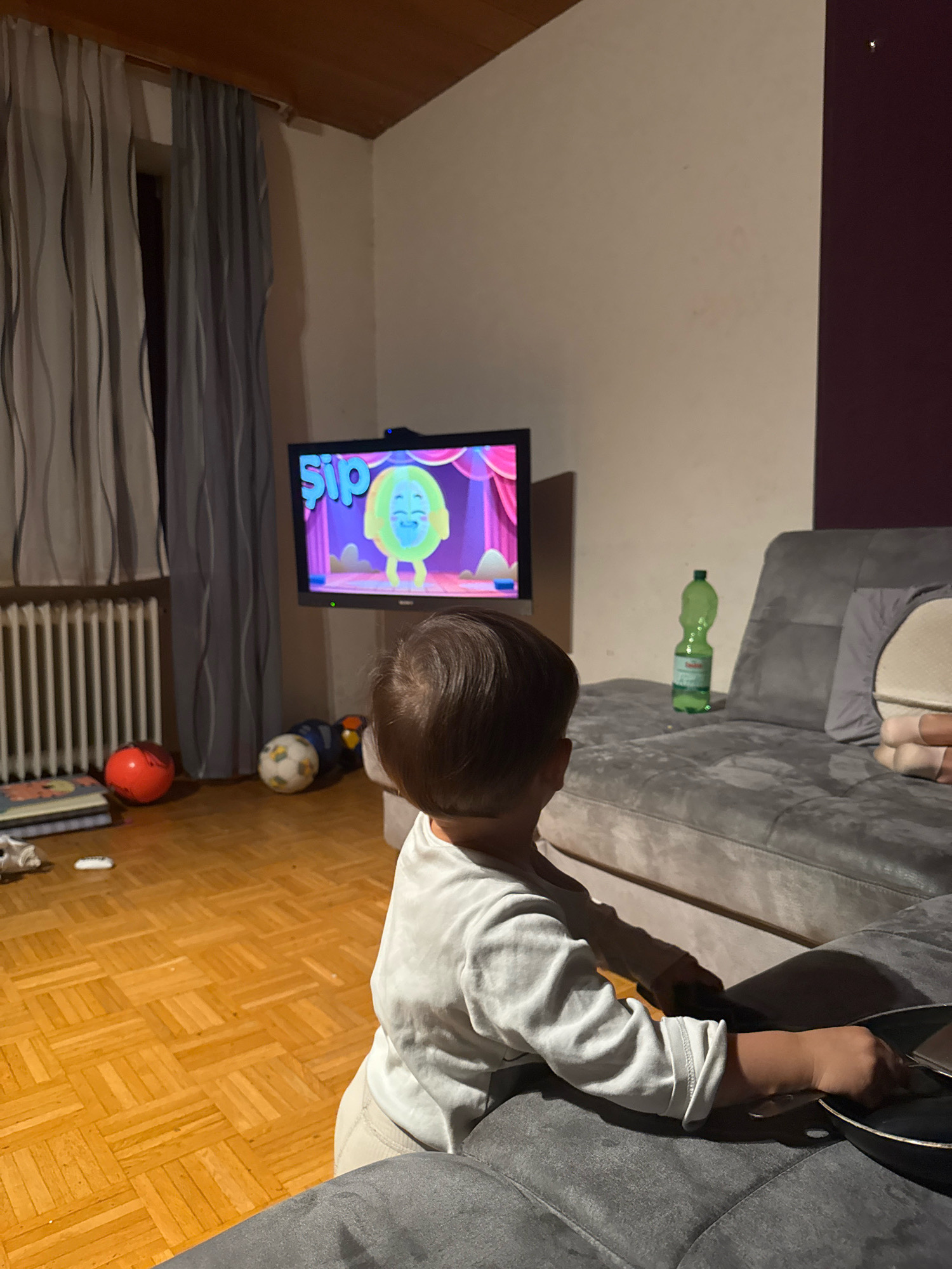 A young child, viewed from behind, is sitting on a couch and watching a television screen in a dimly lit room. The child is wearing a light-colored long-sleeved shirt and beige pants. The television displays a cartoon with a bright green character. In the background, there are curtains, a radiator, several balls on a wooden floor, and a green plastic bottle on the couch. The room appears to be a living room. The presence of a child watching television suggests a domestic setting. The overall ambiance is calm and focused on the entertainment. No specific landmarks or distinguishing features of Überherrn, Germany, are visible in this image.