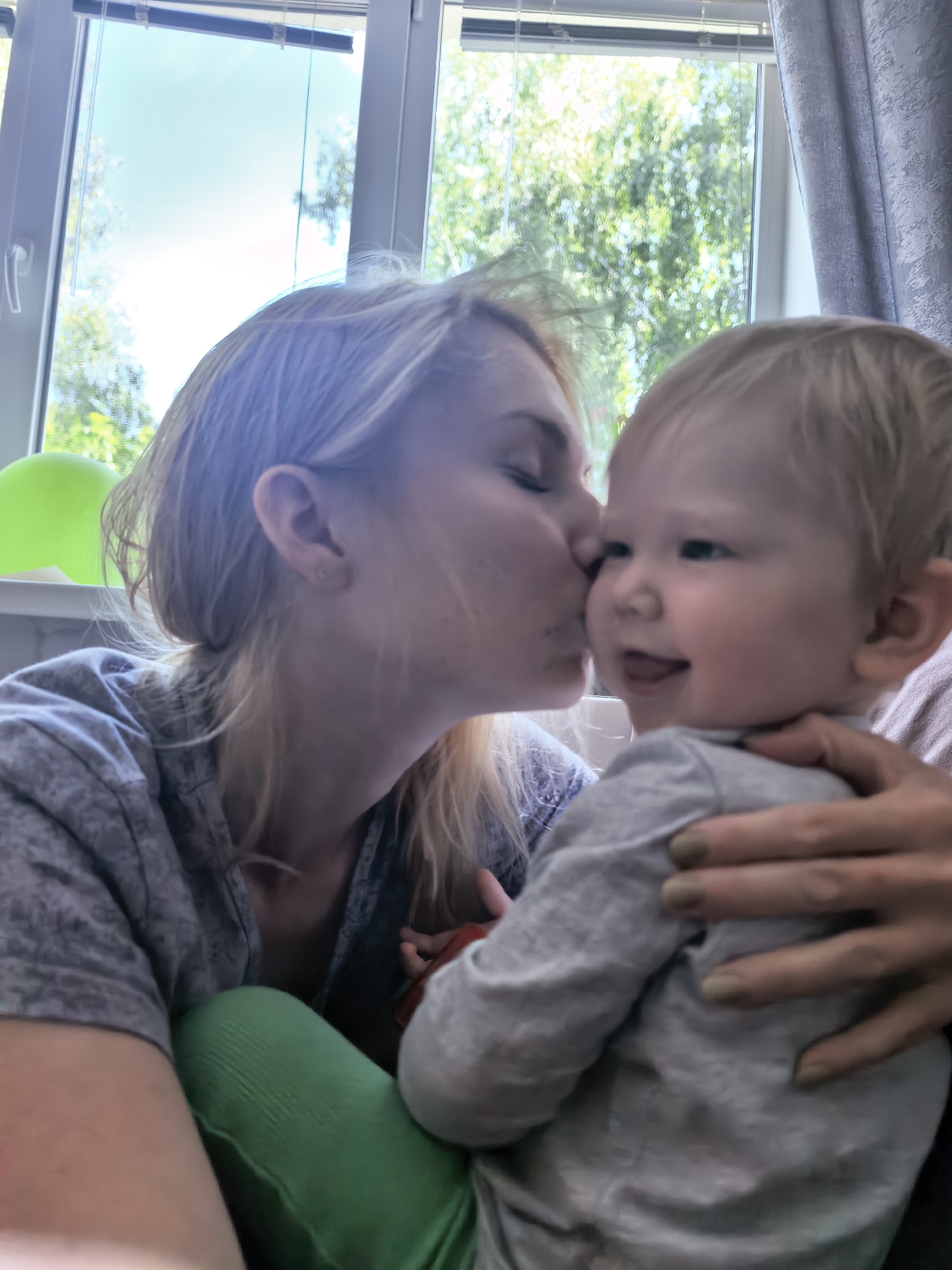 A blonde woman is kissing a baby in front of a window. The woman is wearing a grey shirt and green pants. The baby is wearing a grey shirt. The window has blinds and trees are visible outside. There is a green balloon to the left of the frame.