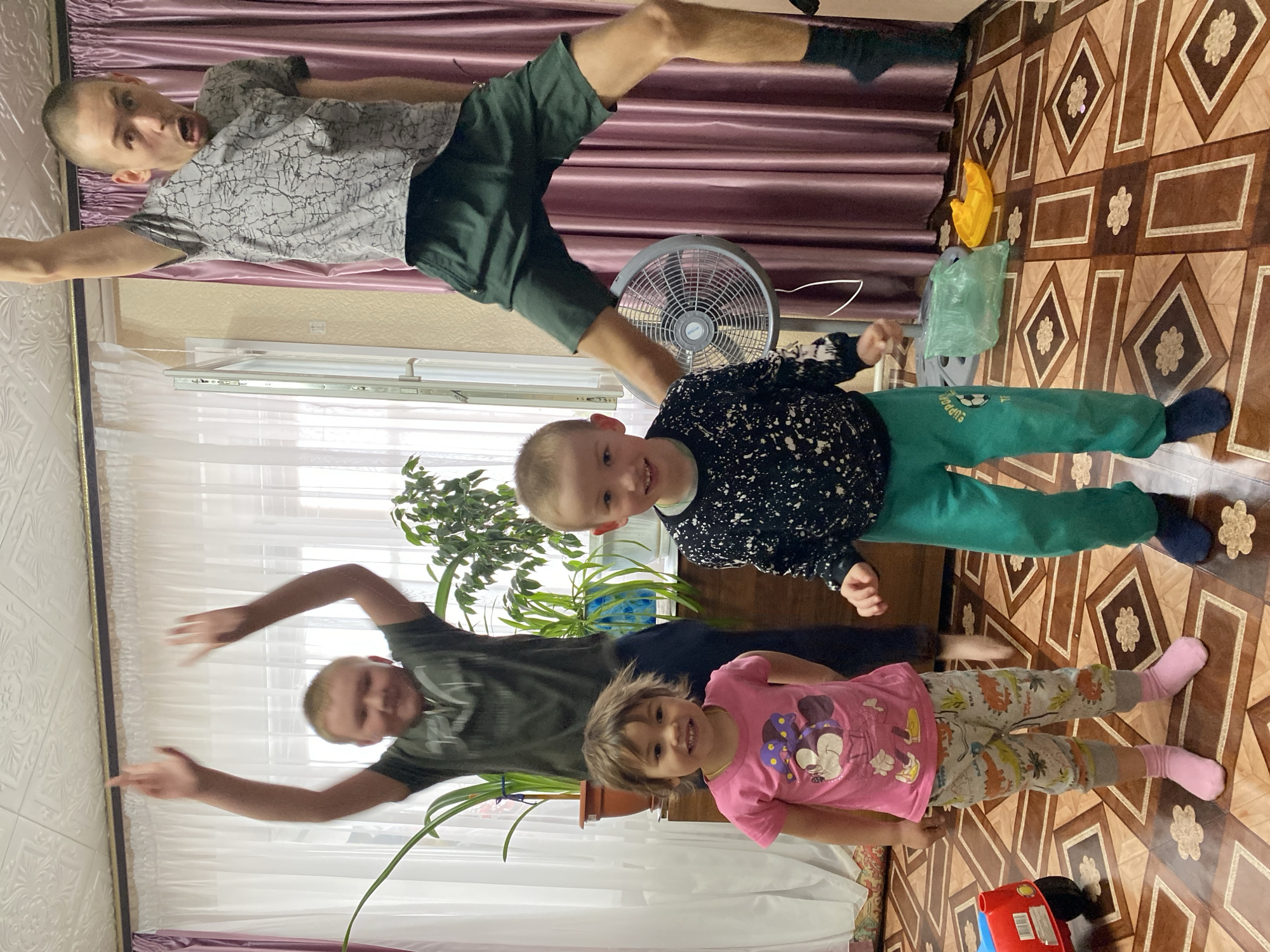 The image depicts four people in what appears to be an indoor setting.
At the top, a young man is lying on his back in the air, his body arched, wearing a patterned gray shirt and dark green shorts. He is wearing black socks.
Below him and more central in the frame, a younger boy is standing. He is wearing a speckled black shirt and green pants. Behind him is an electric fan.
Further down, an older man, is standing with his arms outstretched. He is wearing a dark green t-shirt.
A girl wearing a pink shirt with a cartoon character and patterned pants is standing next to the older man.
Several houseplants are visible in the background.
The floor has a brown patterned design. The wall is covered by purple drapes. A red toy can be seen in the bottom of the image.
There are no obvious clues to determine if this image was taken in Bely Klyuch, Russia.