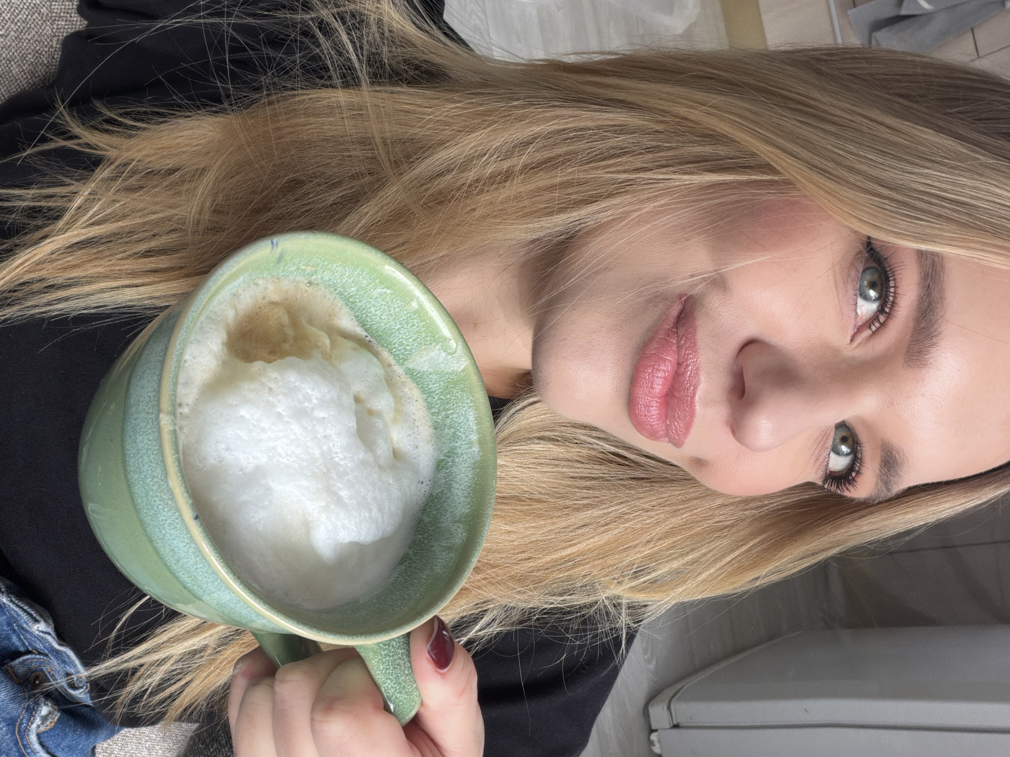 A woman is holding a green mug filled with a foamy beverage, likely a latte or cappuccino. She has light blonde hair, fair skin, and is wearing makeup. She appears to be inside, sitting possibly on a couch, as a hint of blue denim fabric and a dark-colored garment (likely a shirt or jacket) are visible. The backdrop is blurred, but what looks like a white appliance or cabinet can be seen. Without additional clues, specifying this image's location is beyond the scope of information provided.