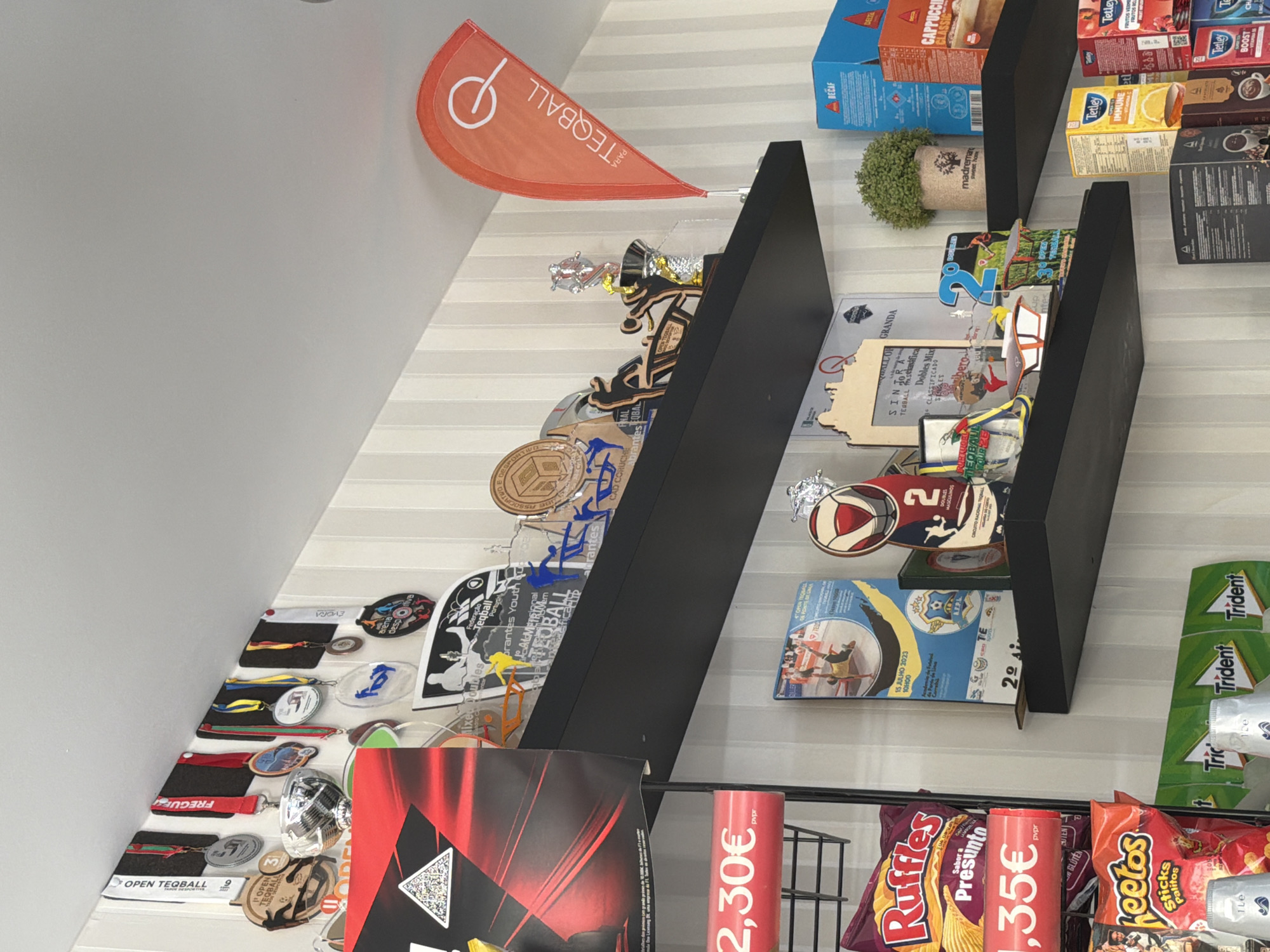 The image depicts an indoor commercial or club setting, likely a cafe or shop associated with the sport of Teqball, located in the Santarém region of Portugal.
The upper section of the scene features a white ceiling and walls covered with light-colored, vertically striped wallpaper. Three black floating shelves span horizontally across the wall.
On the top shelf, numerous medals, some labeled "OPEN TEQBALL" and "FREGUESIA," hang from black felt strips. Various acrylic and wooden trophies and plaques are displayed, many featuring stylized figures of Teqball players. Text on these awards includes "TEQBALL," "Federação Teqball Portugal," and mentions specific Portuguese locations such as "Santarém," "Abrantes," "Almeirim," "Sintra," and "Cad Coruche." A small, orange teardrop-shaped flag with the "TEQBALL" logo stands on the right side of this shelf. A red and black poster with a QR code is partially visible on the far left.
The middle shelf showcases additional Teqball awards, including plaques for "SINTRA" and "Dobles Mix," and a "2º Doubles" award. A small potted artificial plant labeled "madremaria sweet home" sits on the right, next to retail boxes of Delta Decaf coffee and Cappuccino Classic.
The bottom portion of the image displays retail products on a black wire rack. Bags of Ruffles (Sabor a Presunto) and Cheetos (Sticks Palitos) are visible with red price tags showing "2,30€" and "1,35€." Below these, rolls of Trident gum are neatly stacked. On the far right, additional retail boxes of Tetley tea (Immune, Boost, Fruity Infusion) and various coffee products are arranged.
No people are visible in the scene. The items displayed suggest an establishment with a strong connection to Teqball events and achievements within the Santarém district and surrounding areas of Portugal, possibly functioning as a local sports club, academy, or a cafe/shop that celebrates and supports the sport.