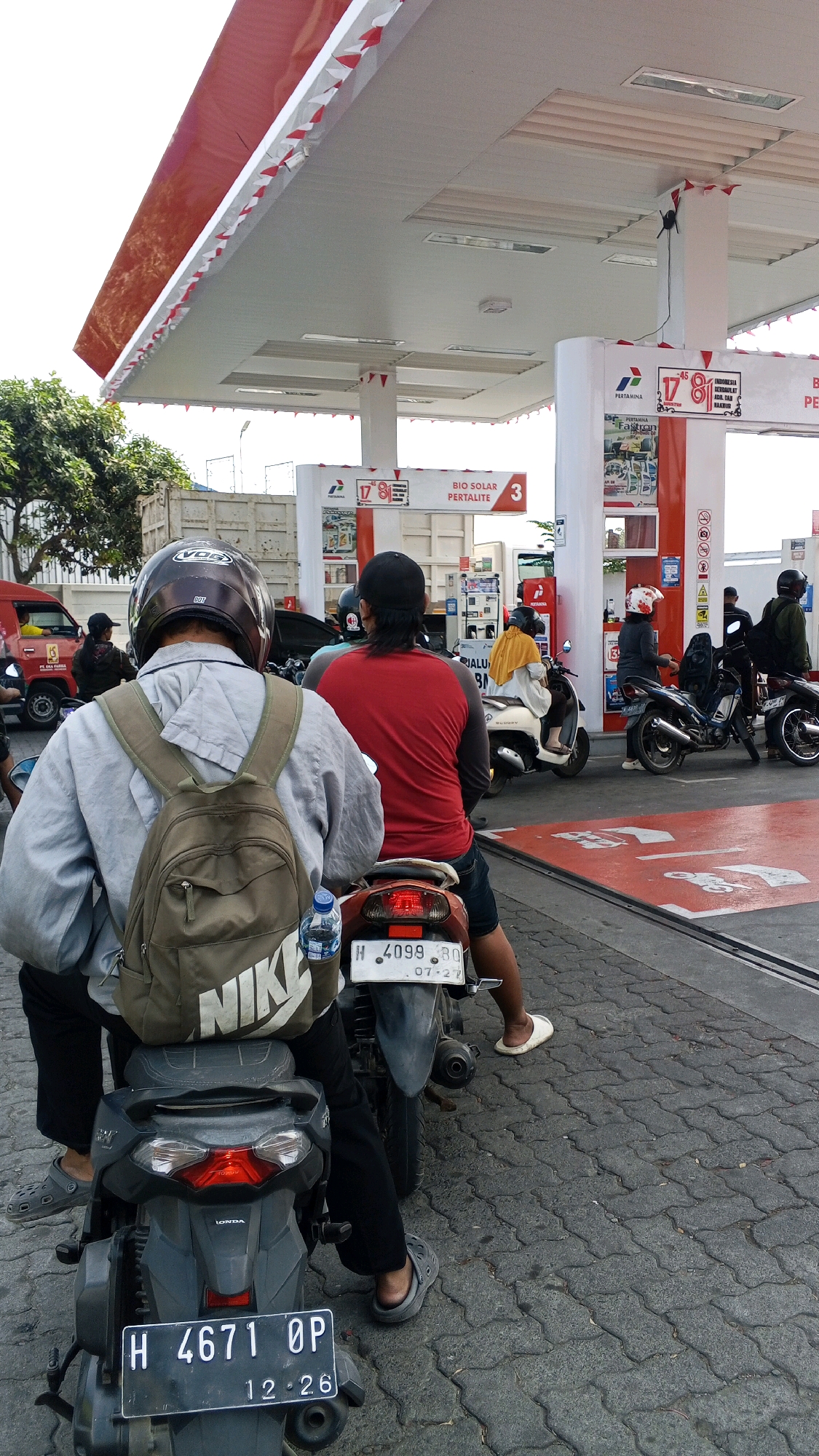 The image shows a street scene in front of a Pertamina gas station in Semarang, Indonesia. Several people on motorcycles are lined up, waiting to purchase fuel. In the foreground, a person wearing a gray long-sleeved shirt, black pants, gray Crocs-style shoes, and a dark brown helmet is seated on a dark gray motorcycle. A khaki backpack with a prominent "NIKE" logo is strapped to their back. A bottle of water is attached to the back of the motorcycle. The motorcycle's license plate reads "H 4671 OP".
Behind this individual, another person wearing a red long-sleeved shirt and black shorts is seated on a maroon motorcycle, also wearing a black baseball cap. Further back, other individuals on motorcycles are visible, including one wearing a yellow headscarf and another person in a black helmet.
The gas station features red and white awnings supported by white pillars. Signs indicate "PERTAMINA" and fuel types such as "BIO SOLAR PERTALITE". The ground is paved with interlocking gray stones, and red painted lines mark lanes. In the background, trees, a white truck, and other vehicles are partially visible. The sky is bright, suggesting daylight.