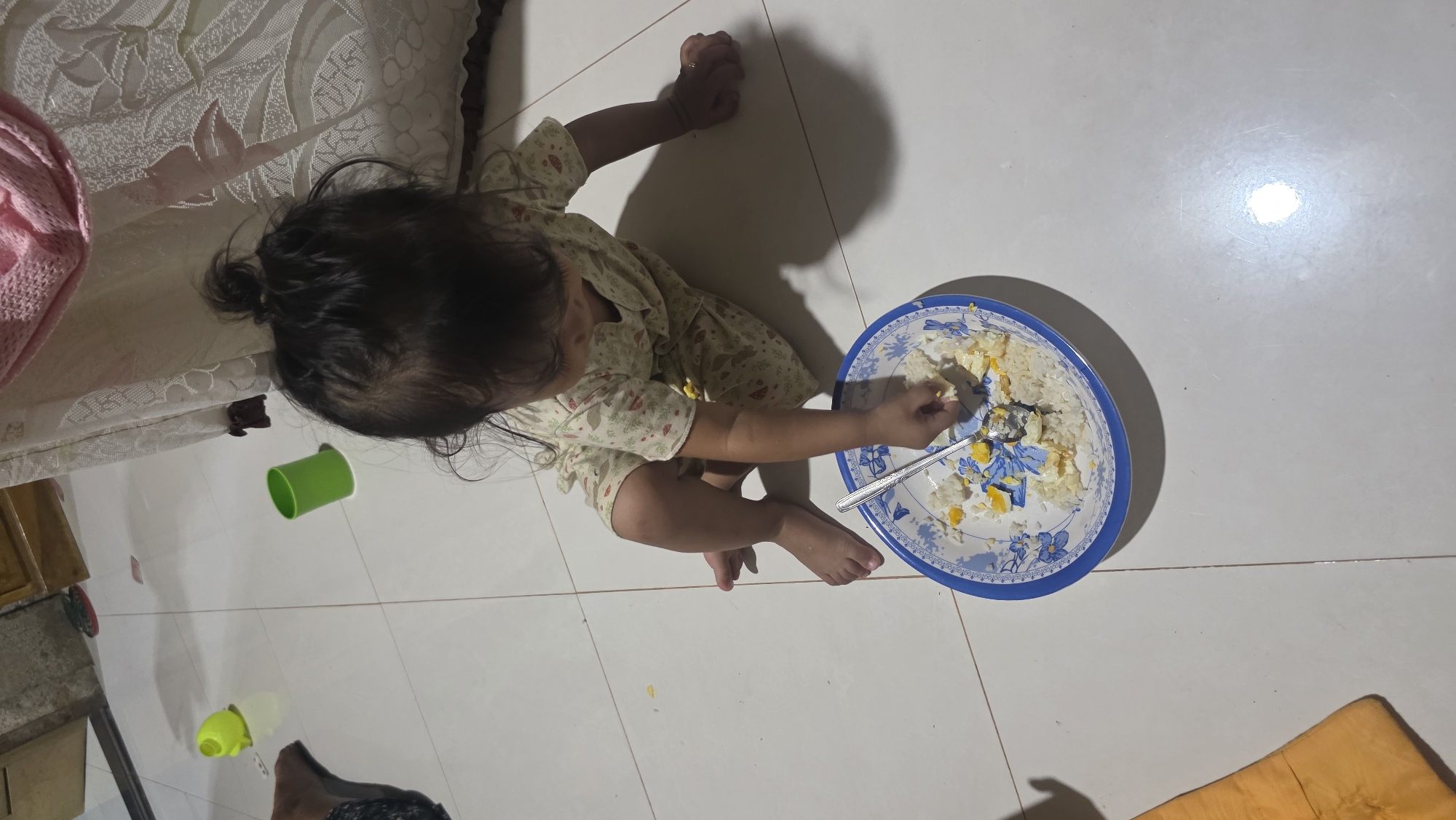 A toddler in a light-colored floral-print outfit sits on a white tiled floor, facing a plate with rice and what appears to be fried egg. She is using her right hand to pick up a piece of food from the plate, while a spoon rests on the plate as well. There are two small plastic cups, one green and one yellow, nearby on the floor. The scene is indoors, with the tile suggesting a domestic setting in Lhokseumawe, Indonesia. A partial view of a couch with a patterned cover and a pink item is visible in the upper left.
