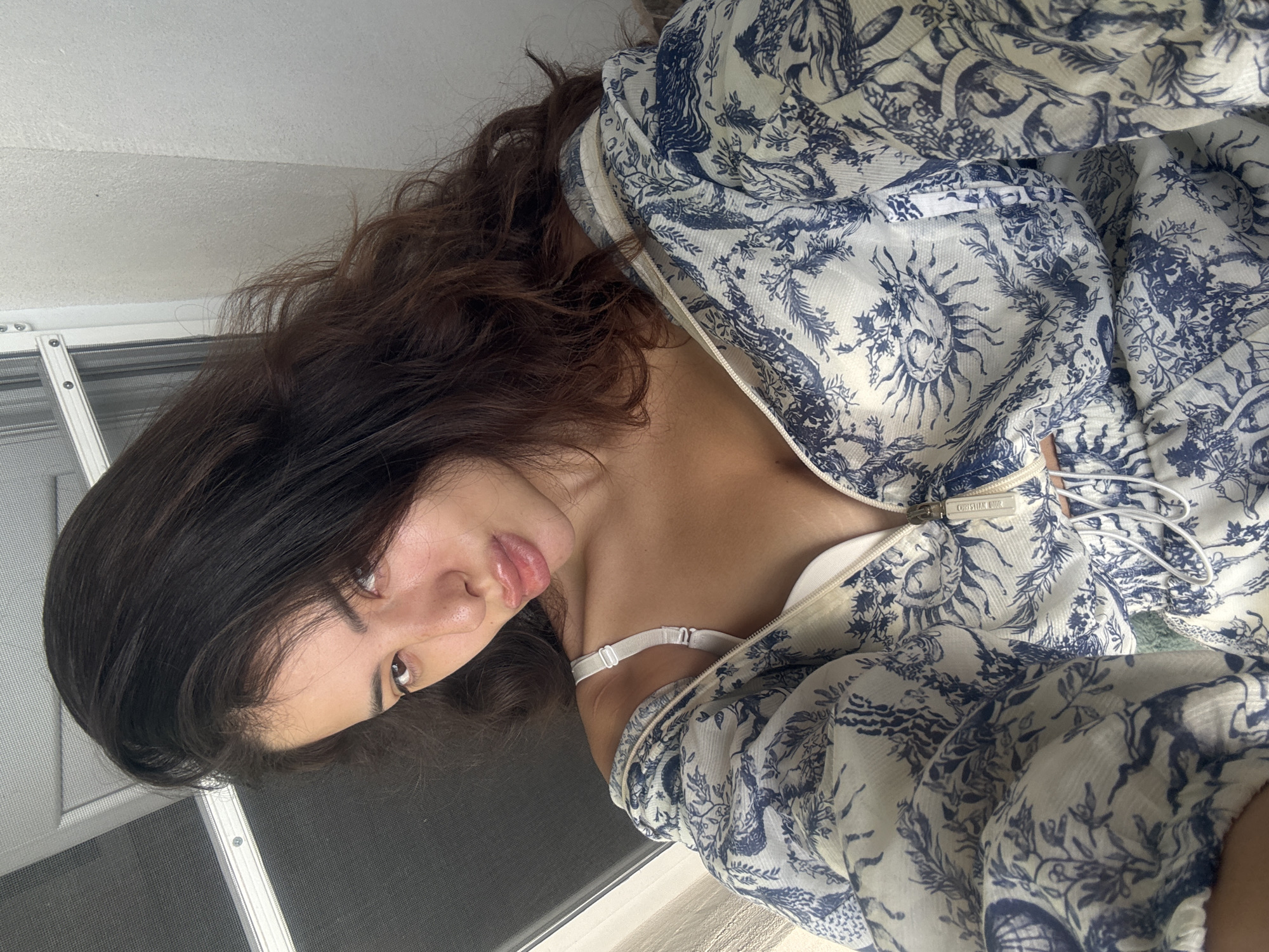 A young woman with long, dark, wavy hair and full lips is shown in a close-up, chest-up selfie. She is wearing a white bra and a white and blue patterned zip-up jacket. The jacket has a toile-style print featuring celestial imagery and various figures. The background appears to be indoors, with a window frame and a textured wall visible. The lighting suggests it is daytime. There is no identifiable context to place this image in the city of Plovdiv, Bulgaria.