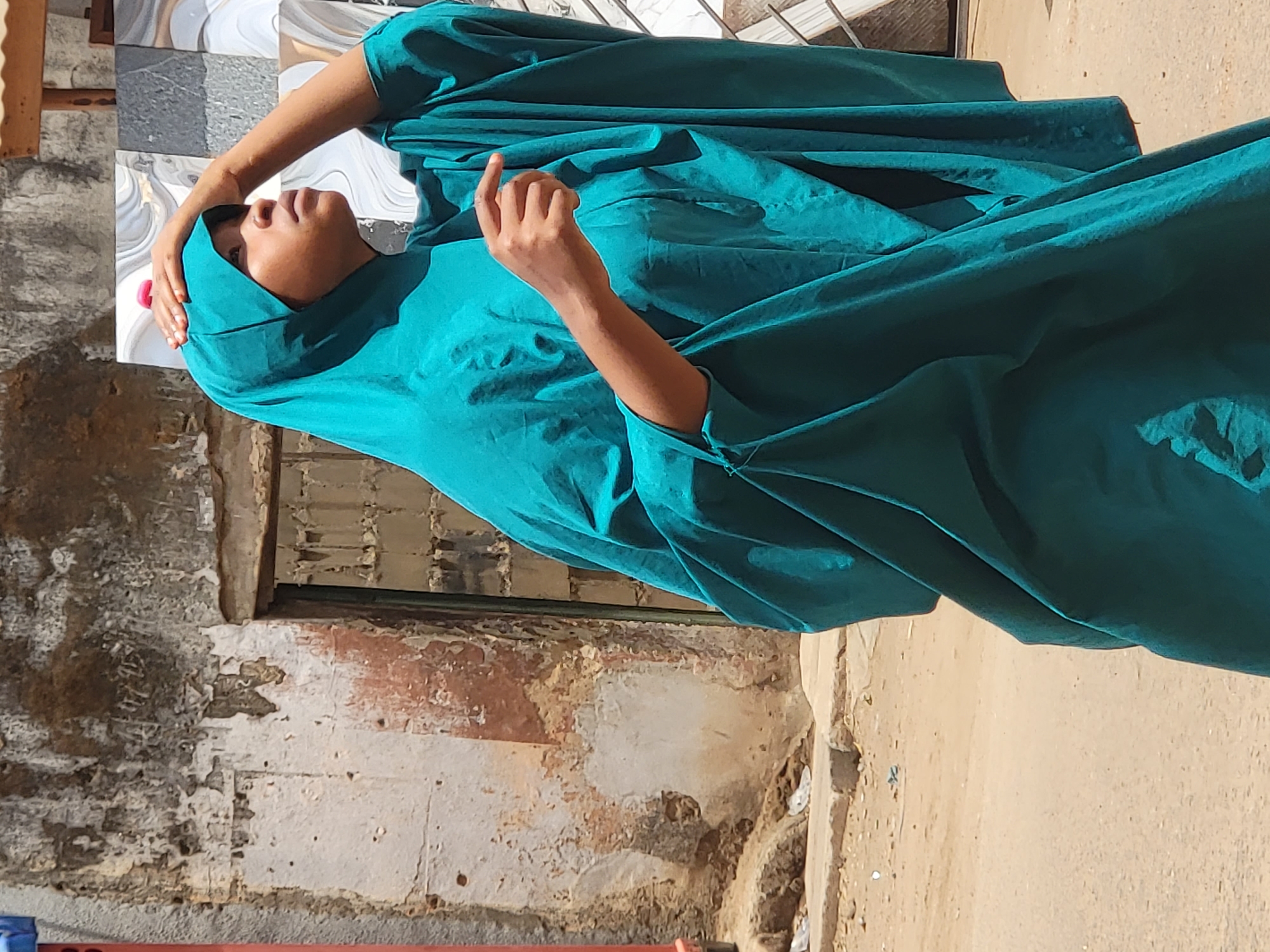 The image shows a person wearing a turquoise headscarf and draped cloth standing next to a building with deteriorating walls in Maiburiji, Nigeria. The ground is sandy. The person has one hand on their head and the other outstretched. The building's wall shows exposed bricks and peeling paint.
