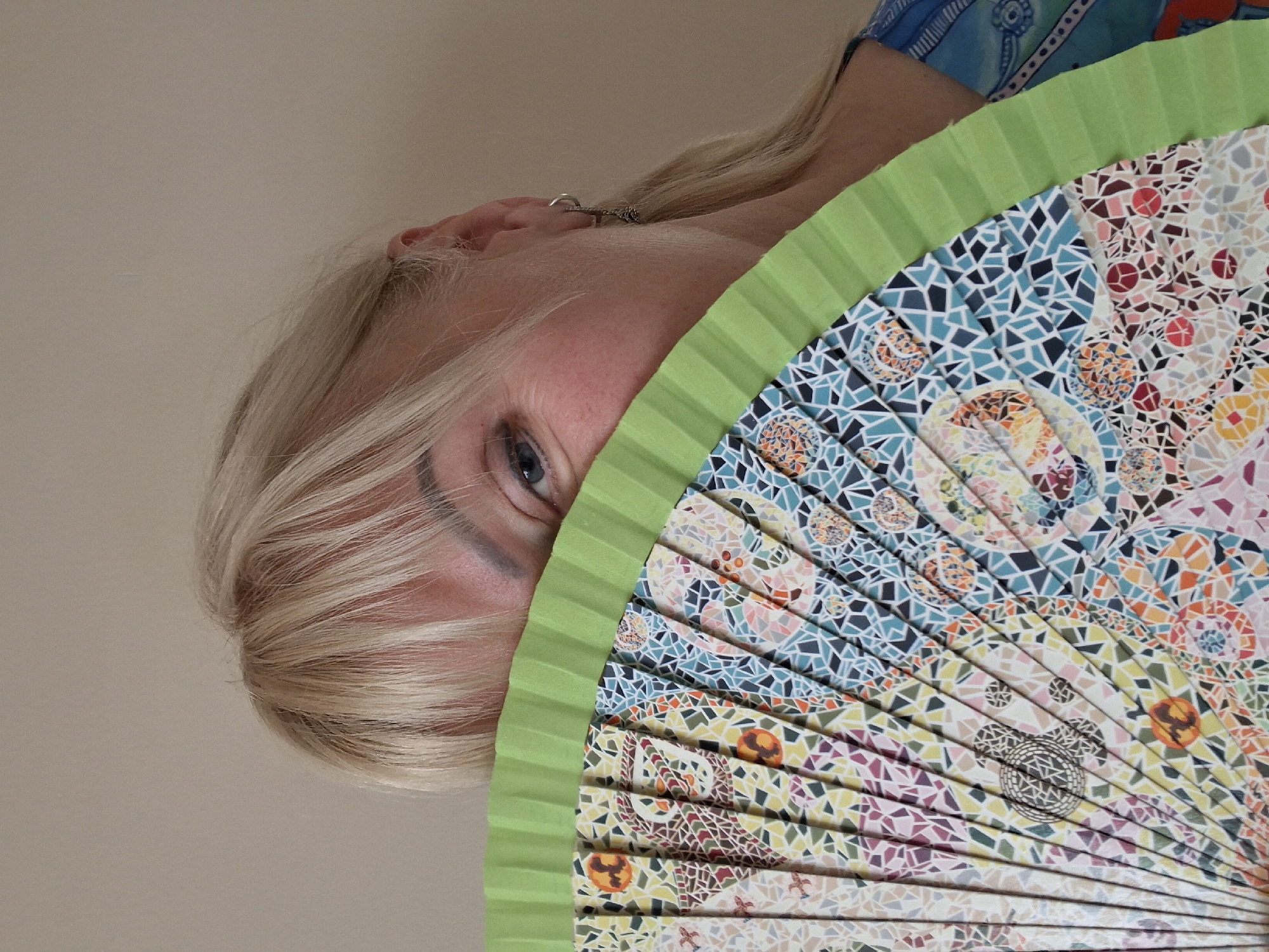 A woman with blonde hair is partially obscuring her face with a colorful fan. She is wearing a blue garment with a patterned neckline. The background is a plain, light-colored wall. The scene appears to be indoors, with neutral lighting. There are no discernible elements that place this image in the city of Alytus, Lithuania.