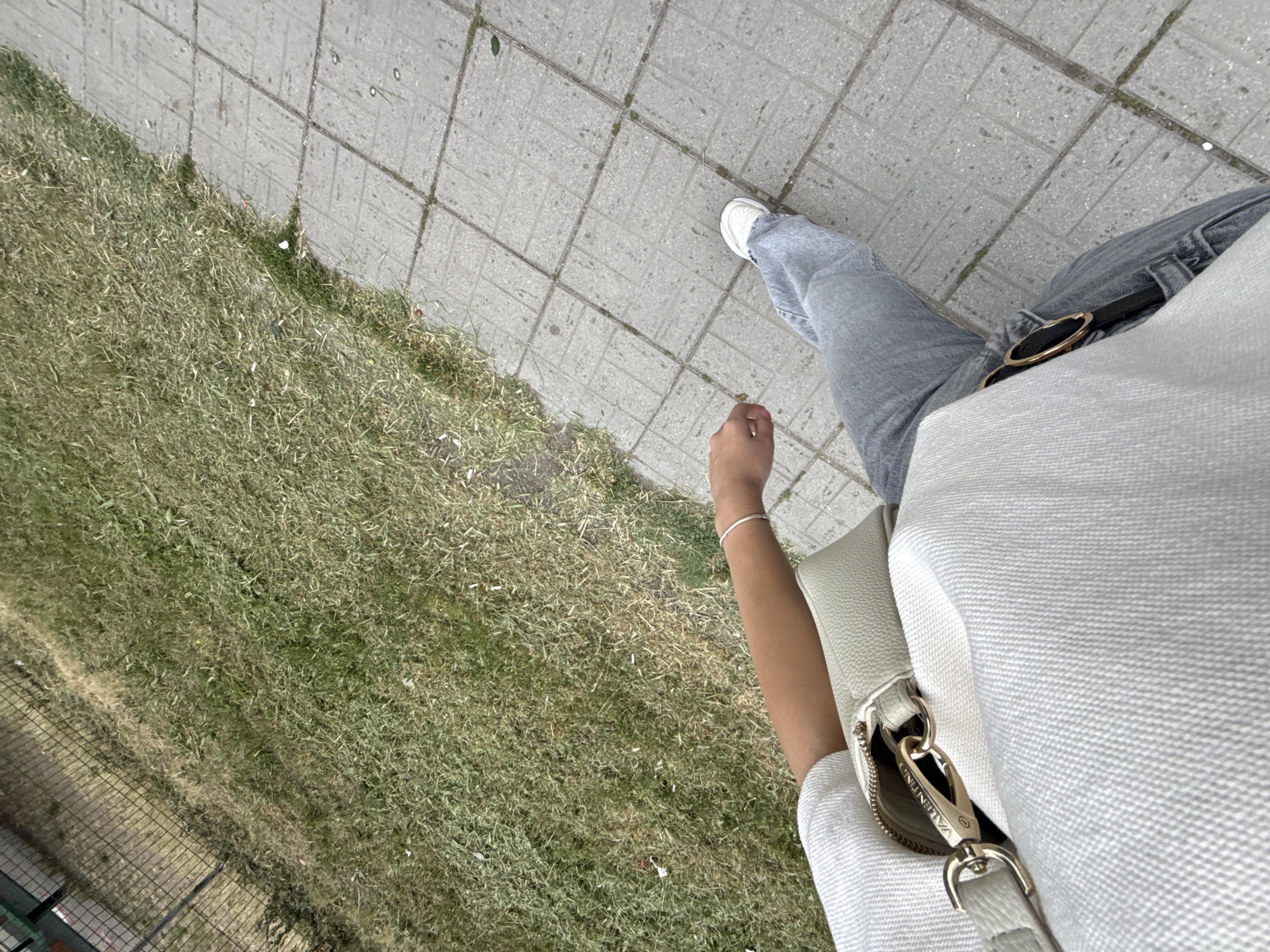The image shows a first-person perspective of a person walking on a sidewalk next to a grassy area. The person is wearing light-colored jeans, a light-colored top, and white sneakers. A light-colored purse is visible on the person's left hip. The sidewalk is made of gray concrete tiles, and the grassy area appears to be a park or a verge next to a street. The scene is outdoors, and the lighting suggests it is daytime. The location is identified as Sofia, Bulgaria.