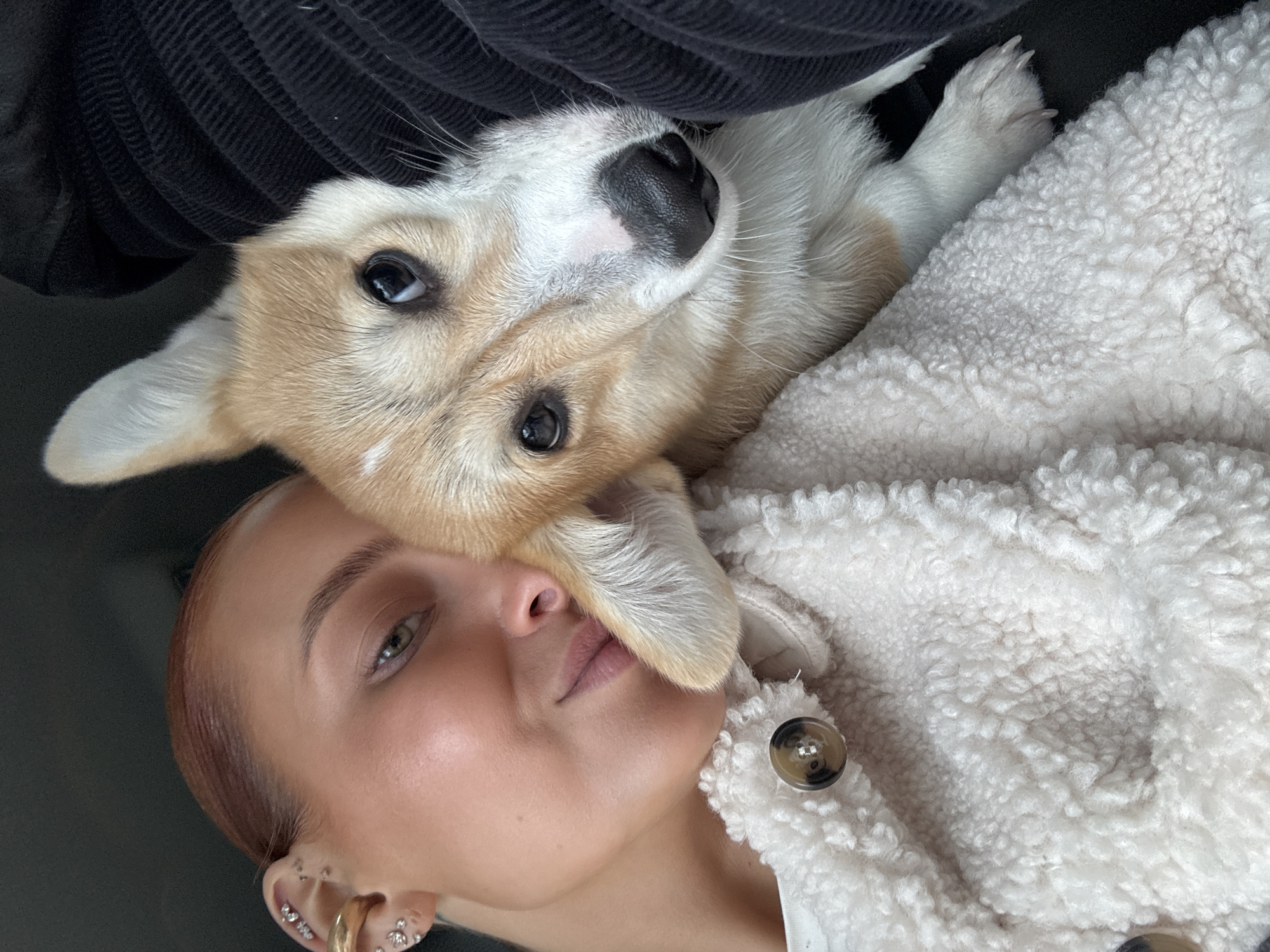 A young woman with short, light brown hair and multiple piercings in her left ear is lying down. A tan and white Pembroke Welsh Corgi puppy is lying on top of her chest, with its head and paws resting on her face. The woman is wearing a white, fleece jacket with brown buttons. A dark, corduroy material is visible behind the dog's head, possibly a piece of clothing or furniture. No information is available to verify the location is Chaikovsky, Russia.