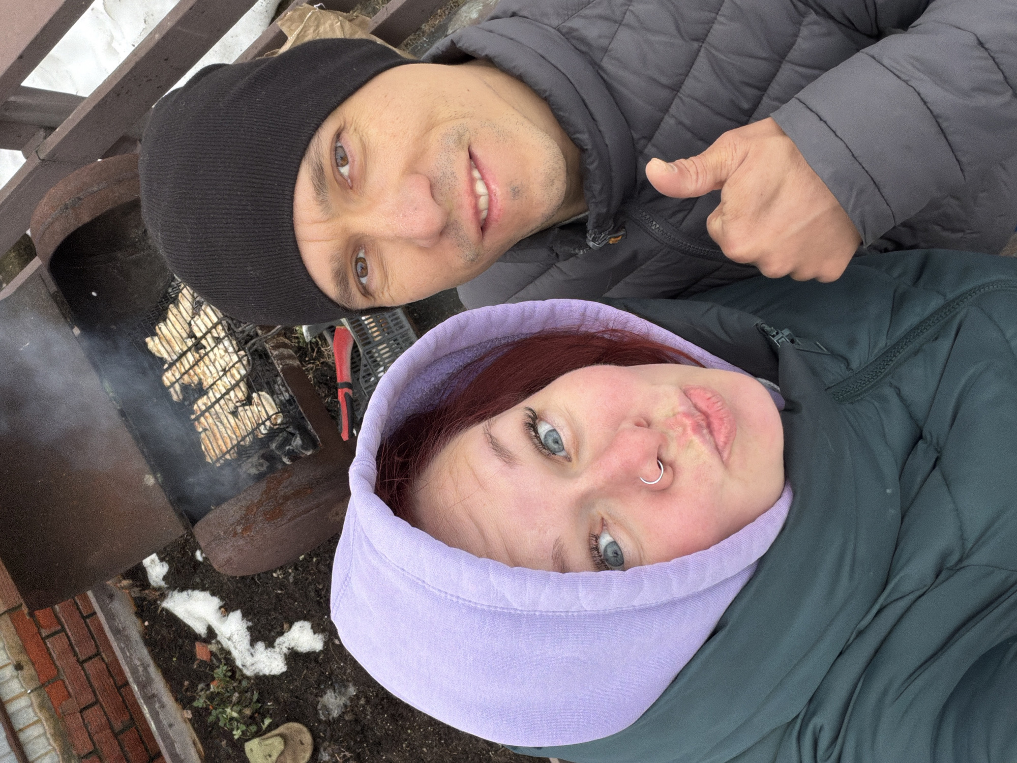 Two people, a man and a woman, are posing for a selfie. The man is wearing a black beanie and a gray quilted jacket and gives a thumbs up. The woman is wearing a lavender hood and a dark green jacket. Behind them is a barbecue grill with food cooking over a fire. Snow is on the ground. The scene appears to be outdoors near a brick wall. The location is in or near Chaikovsky, Russia.