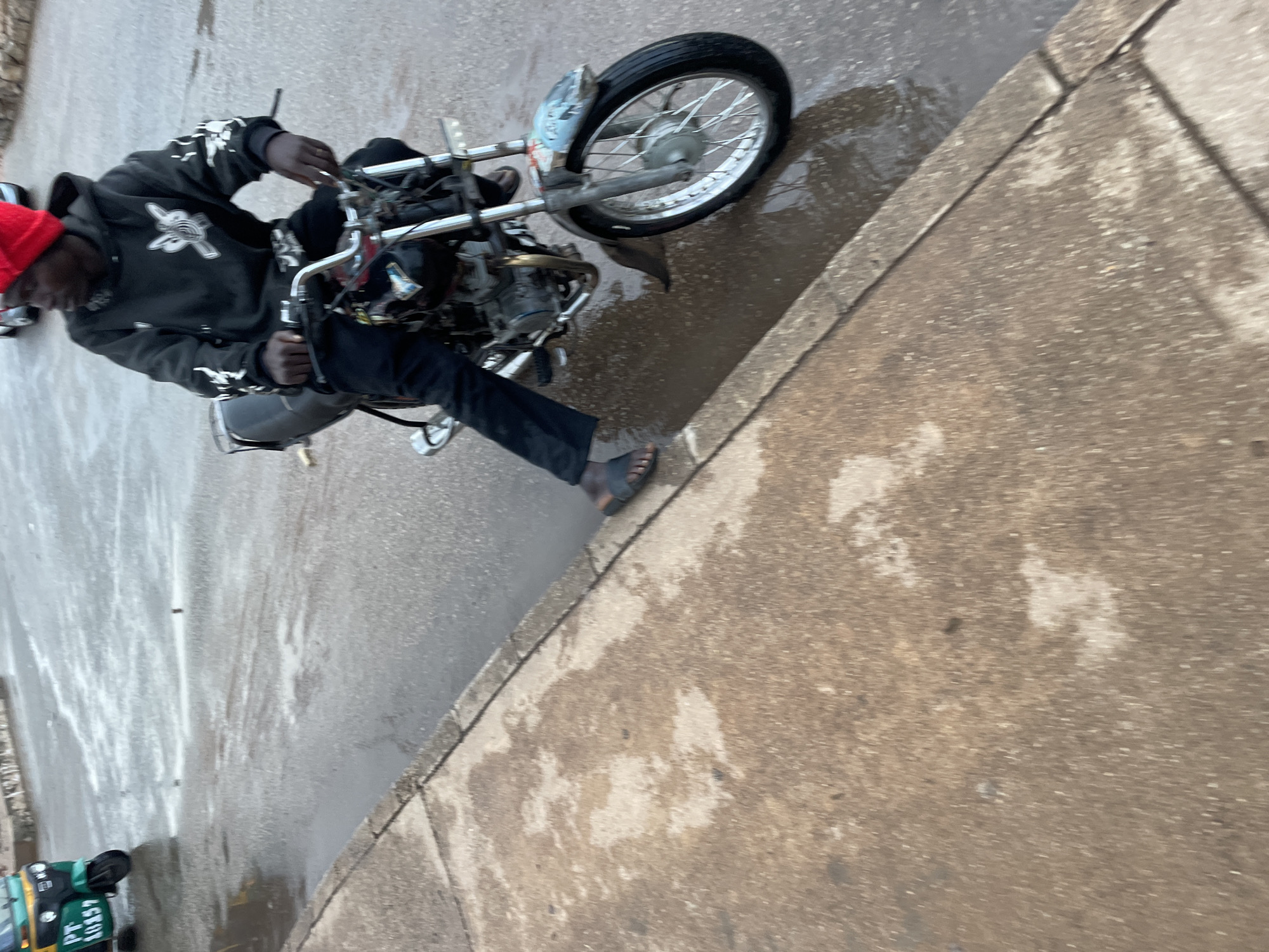 A person wearing a red beanie and a black hoodie with white graphics is riding a motorcycle on a wet, asphalt street in Jos, Nigeria. The motorcycle is dark colored and appears to be a custom or modified model. The rider is wearing dark pants and sandals. In the background, a green auto-rickshaw with the license plate "PT 10157" is visible. The street is covered in puddles and reflections, indicating recent rain. The sidewalk, made of concrete, is also wet. The overall scene suggests a cloudy or overcast day.