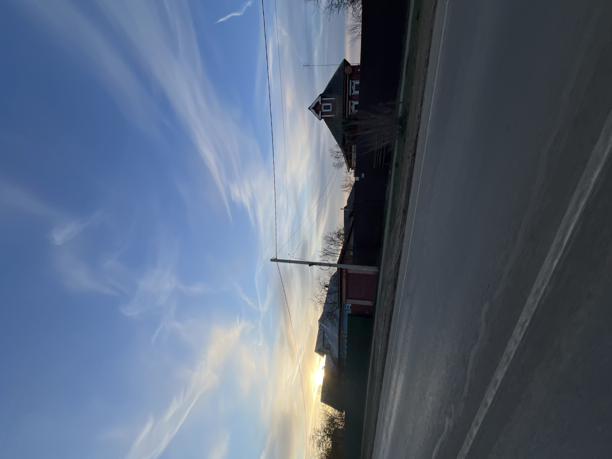 The image presents a roadside view under a partly cloudy sky. A road runs along the right side of the frame. Adjacent to the road, a row of buildings, including a distinctive house with a gable roof and another building with a red-painted area, are visible. Utility poles and wires stretch across the sky above the buildings. The lighting suggests either sunrise or sunset, with the sun low on the horizon.
The location is not explicitly visible from the photo itself; Yasenevo is mentioned in the prompt but not visually present.