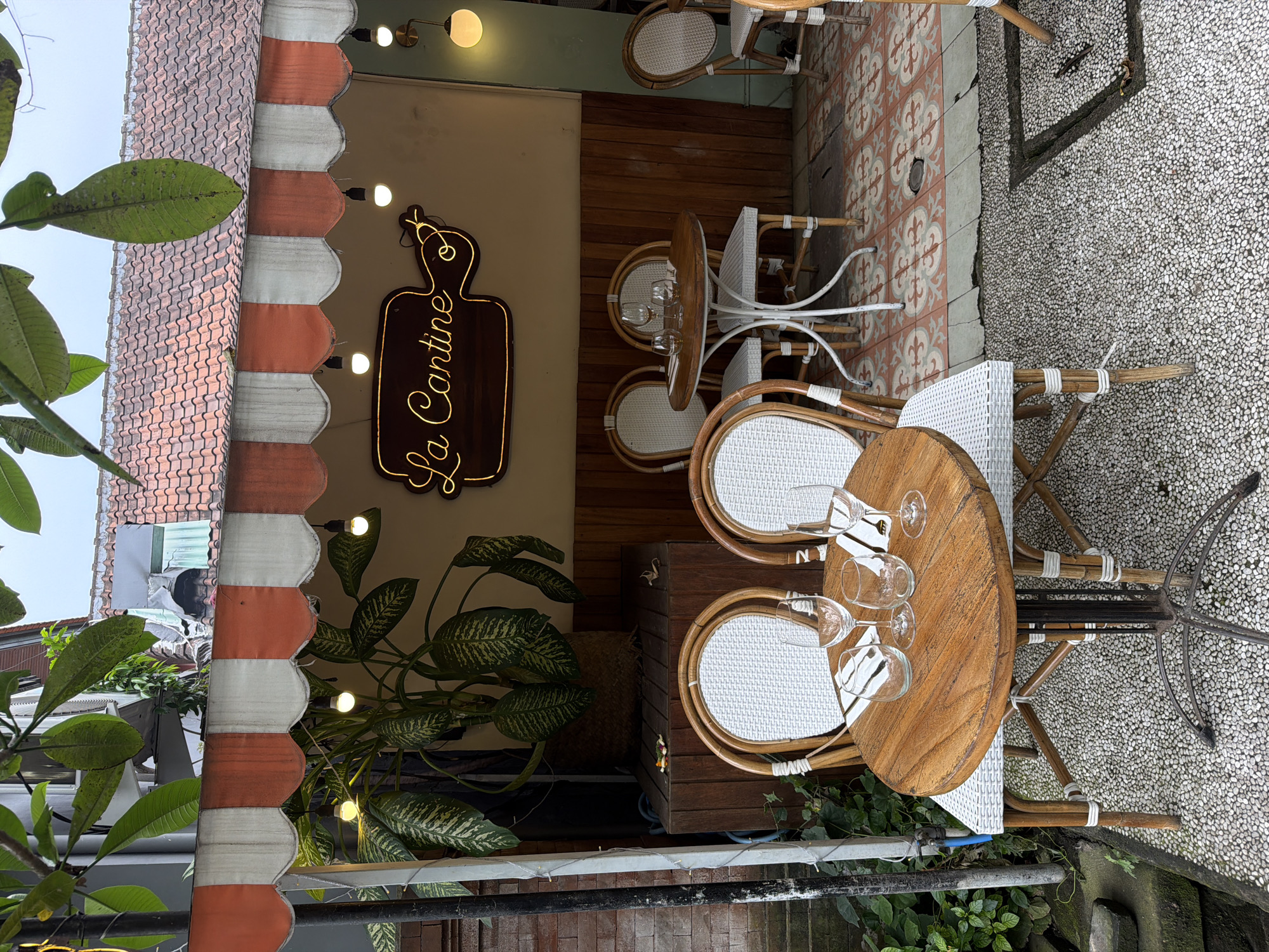 The image presents an inviting outdoor dining area of a cafe or restaurant, likely located in Ubud, Indonesia. The scene is captured with a vertical orientation, showing a blend of natural and crafted elements.
In the foreground, several round wooden tables are arranged, each set with multiple empty wine glasses and white placemats. These tables are accompanied by chairs featuring white wicker-patterned seats and backrests, supported by frames made from rattan and white metal.
A prominent neon sign, shaped like a cutting board, spells out "La Cantine" in luminous cursive script. This sign is mounted on a light beige wall that transitions into horizontal wooden paneling below. Tropical plants with large, variegated green leaves are strategically placed in pots, enhancing the natural aesthetic. Small, spherical light fixtures are visible, both wall-mounted and strung as fairy lights, contributing to a warm ambiance.
Above the main cafe area, a scalloped awning with alternating orange and white stripes provides partial cover. Beyond the awning, traditional terracotta roof tiles and lush green foliage from nearby trees, including distinctive large leaves in the immediate foreground, are visible. In the background, partially obscured, an individual with dark hair and wearing a light garment can be seen near the roofline, though their activity is not discernible.
The flooring transitions from decorative red, white, and grey patterned tiles closer to the cafe to a light-colored pebble wash concrete surface further out. A dark metal railing or fence runs along the bottom edge of the frame. The setting exudes a peaceful, casual, and tropical vibe. The time of day appears to be daytime, possibly late afternoon or an overcast day, given that the artificial lights are on while the sky above is bright.