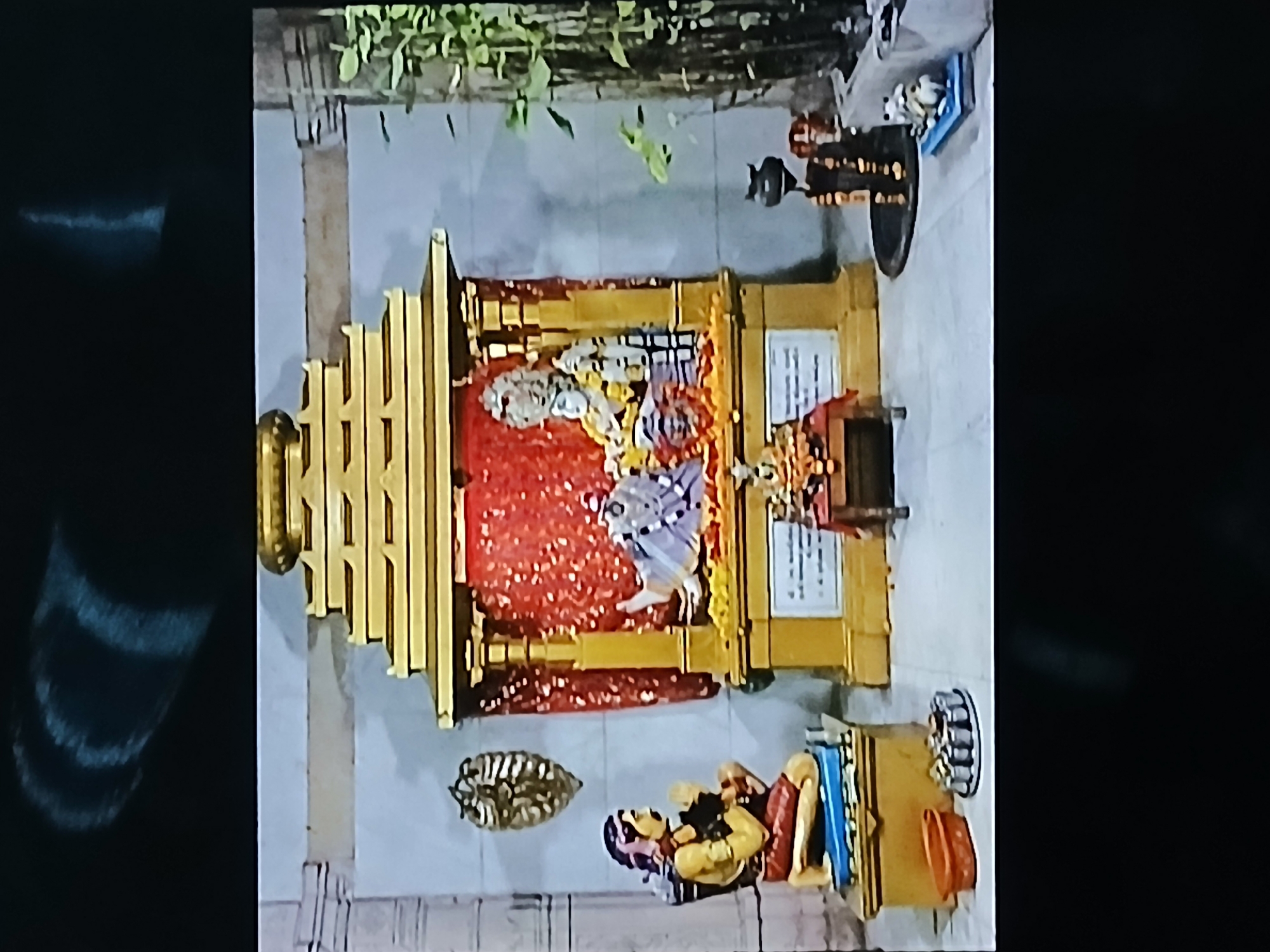The image shows a Hindu temple shrine. The shrine is gold colored with a red, sequined backdrop. Two deities are placed inside the shrine. A figure is seated in front of the shrine, with books and other items placed around it. Other religious items and figures are placed nearby. The location is most likely in a temple in Bengaluru, India, based on the shrine's style and the religious items present.