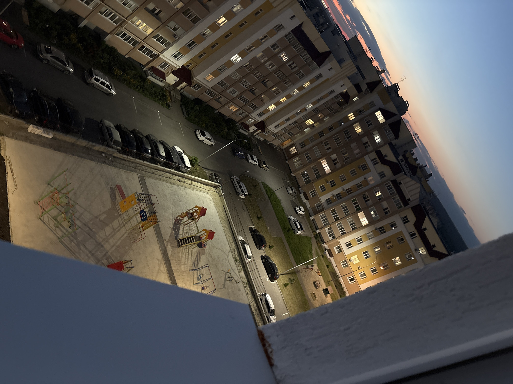 This media file shows an aerial view of an apartment complex and its surroundings. The subjects include multiple buildings with many windows, some of which are lit, indicating residents. There are also several cars parked on the street and in parking areas. A playground with colorful equipment is visible in the foreground.
The scene is set outdoors, likely in a residential area. The time of day appears to be dusk or early evening, as suggested by the soft, warm light illuminating some of the windows and the colors in the sky. The sky displays a gradient of blue and pink hues, characteristic of sunset or sunrise. There are no people actively engaged in any particular activity, and the weather appears calm.
Notable details include the architectural style of the buildings, which seem to be multi-story apartment blocks with balconies. The cars are varied in color and model, suggesting a typical urban or suburban setting. The playground equipment is a prominent feature of the outdoor space. The perspective is from a high vantage point, looking down at the scene. No visible text is present in the image. The overall mood conveyed is one of quiet residential life at the end of the day.