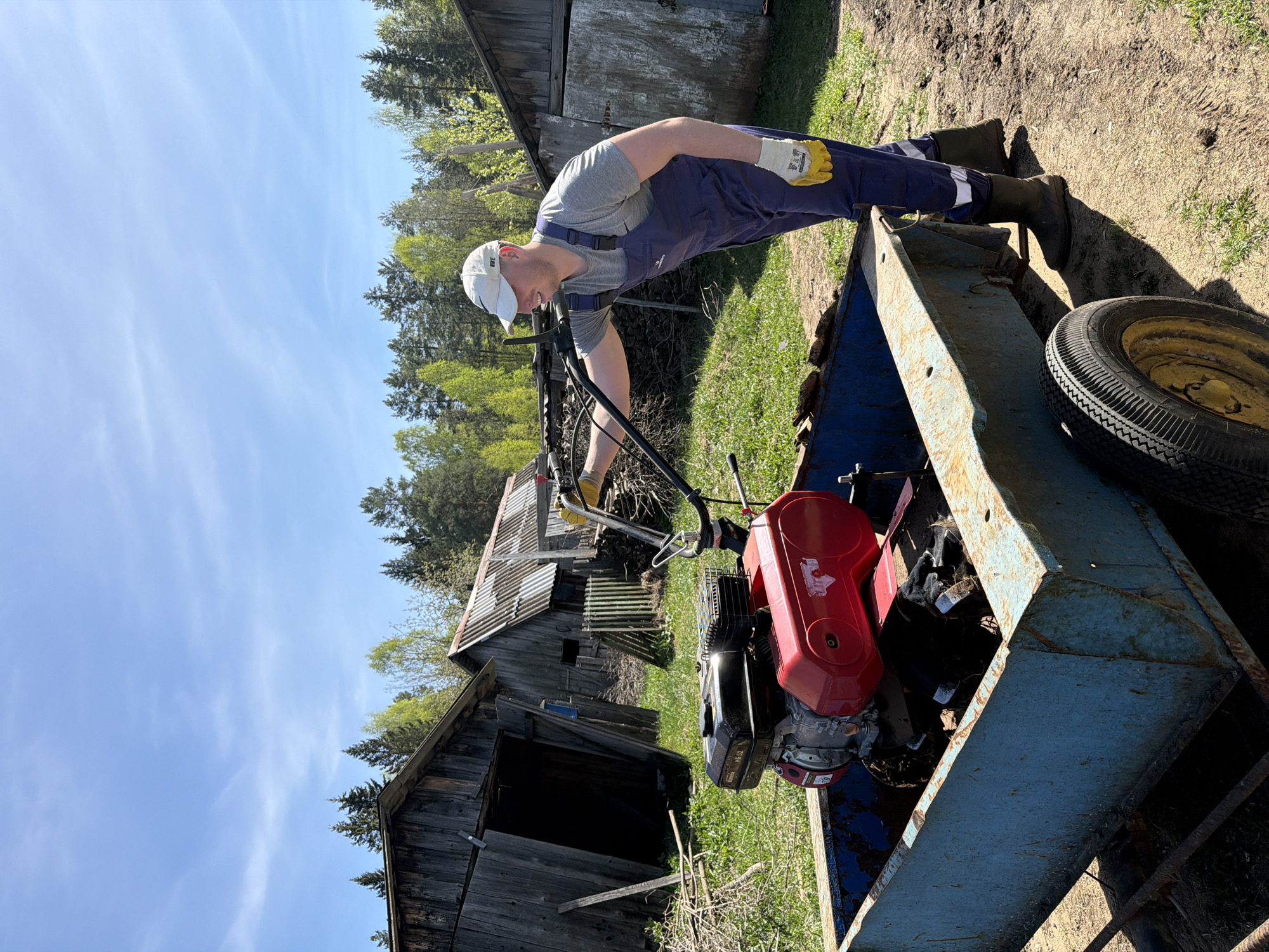 A man wearing blue overalls, a grey t-shirt, a white baseball cap, gloves, and dark rubber boots is leaning over a blue trailer. He is holding the handle of a red and black rotary tiller that is inside the trailer. A yellow and black spare tire is visible in the trailer. Behind the trailer is a grassy area with two weathered wooden buildings, one of which appears to be a barn. Trees are visible in the background, and the sky is blue with some clouds. The location is Уральское, Russia.
