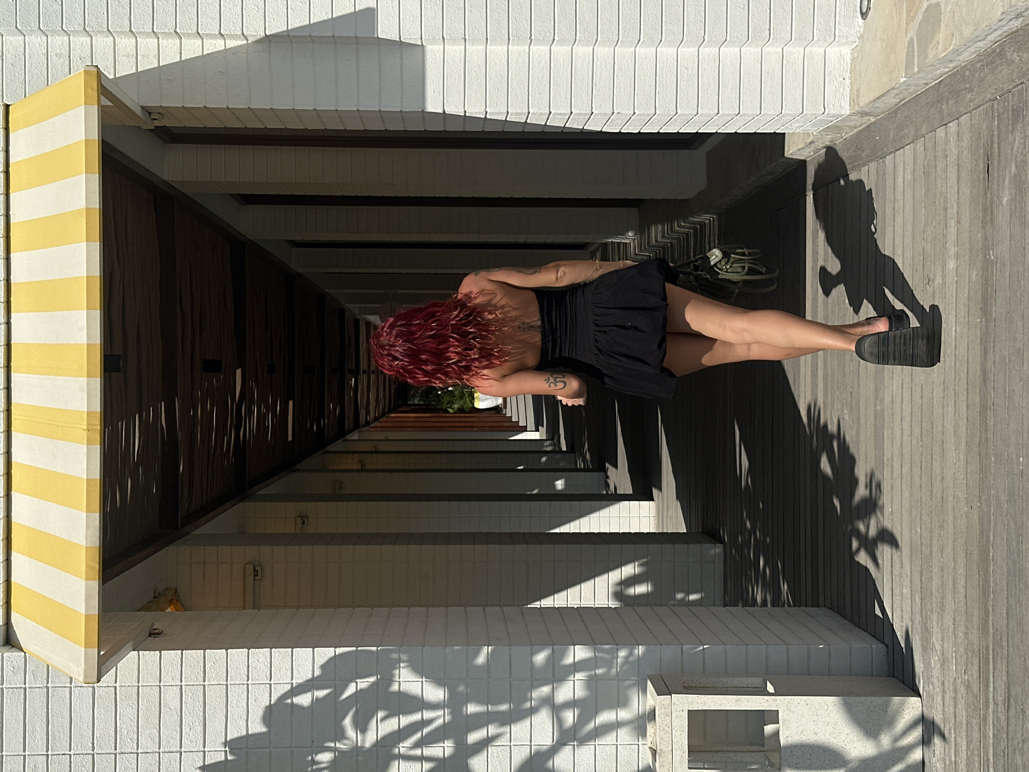 The image captures a woman with red curly hair, wearing a black dress and sandals, standing in a hallway. The hallway has a distinctive architectural design, with white tiled walls and supports interspersed with sections of wooden cladding. A yellow and white striped awning is visible at the hallway entrance. The floor appears to be made of wooden planks. A pair of green sandals is slightly visible in the background. The environment is brightly lit, casting shadows on the floor and walls. The location is presumed to be in the city of Unknown, Indonesia.