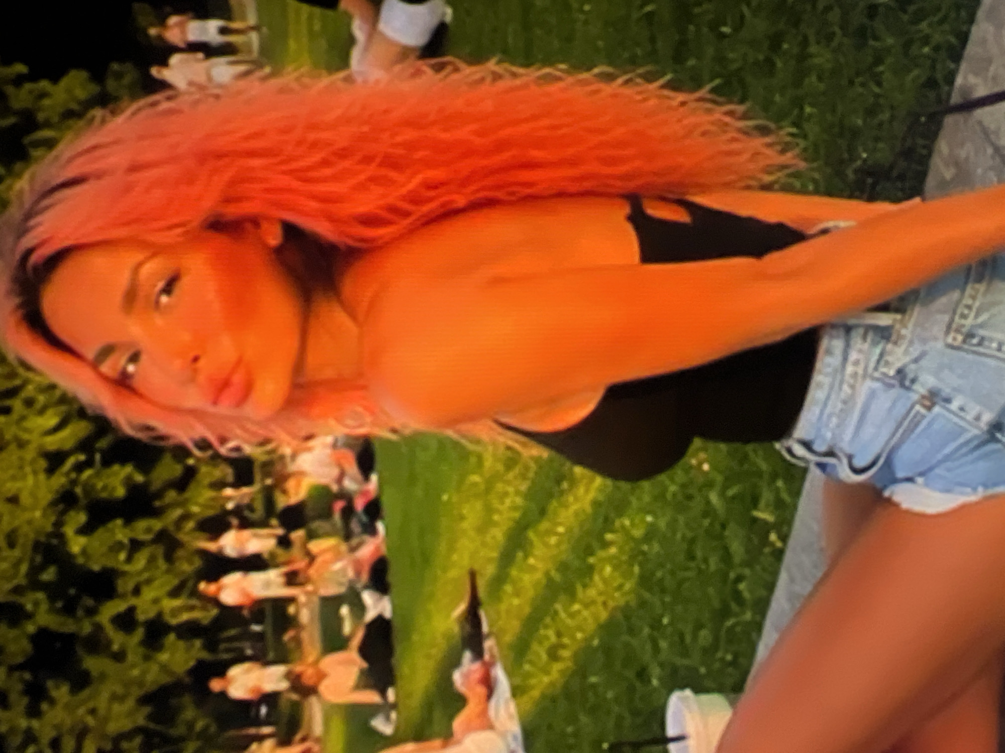 A woman with bright orange hair sits on a stone ledge in an outdoor park at dusk. She is wearing a black tank top and denim shorts. Her face is turned towards the camera, and she is making a kissing gesture. In the background, several other people are seated on the grass, some of whom appear to be watching an event or gathering. The lighting suggests the sun has recently set, casting long shadows across the grass.
