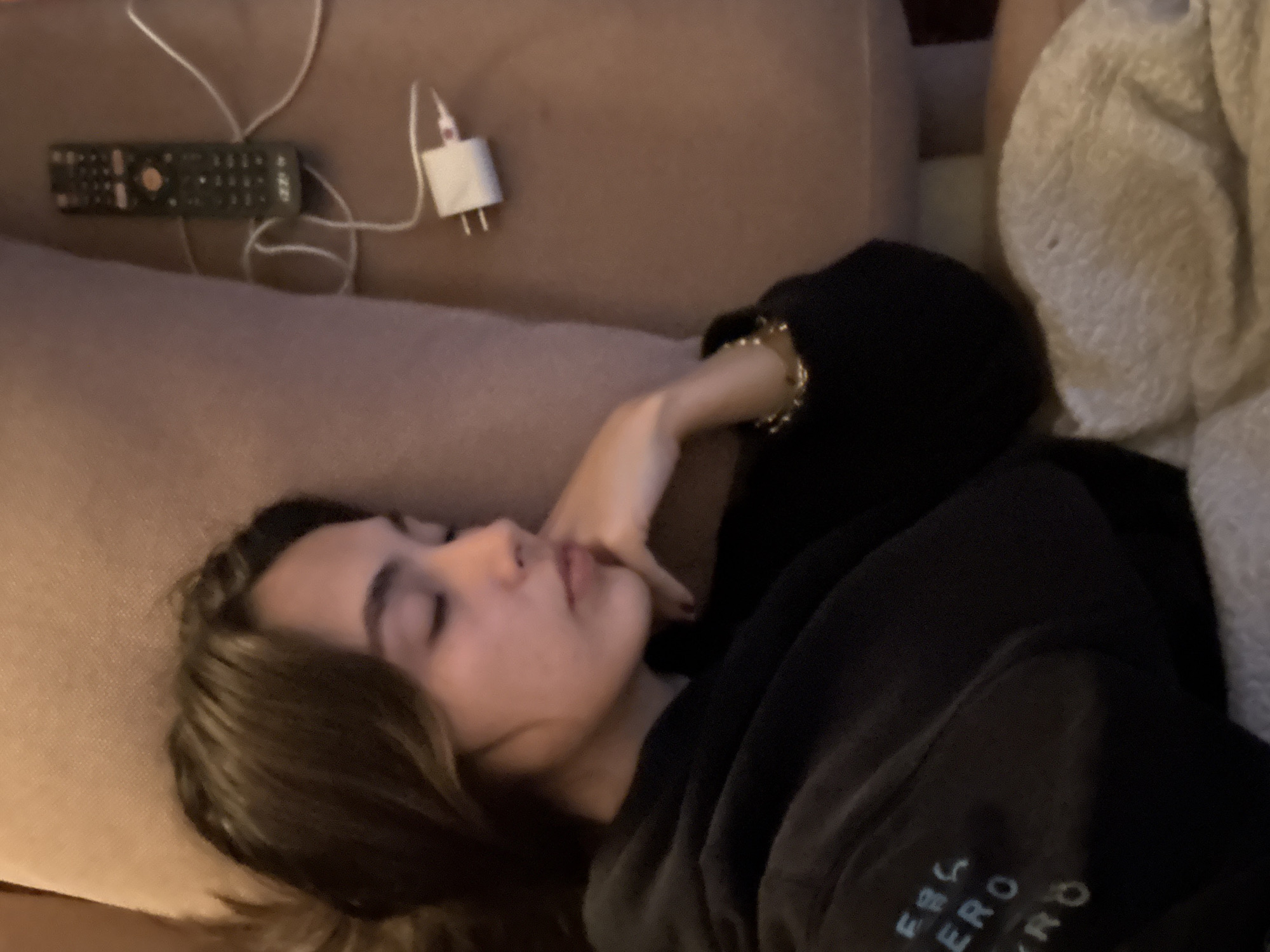 A young woman with dark hair is sleeping on a couch. She is wearing a black hoodie and a gold bracelet. Her hand is resting on her face. On the couch next to her is a remote control and a phone charger. There is also a white blanket.
The location is not identifiable as Mexico City.