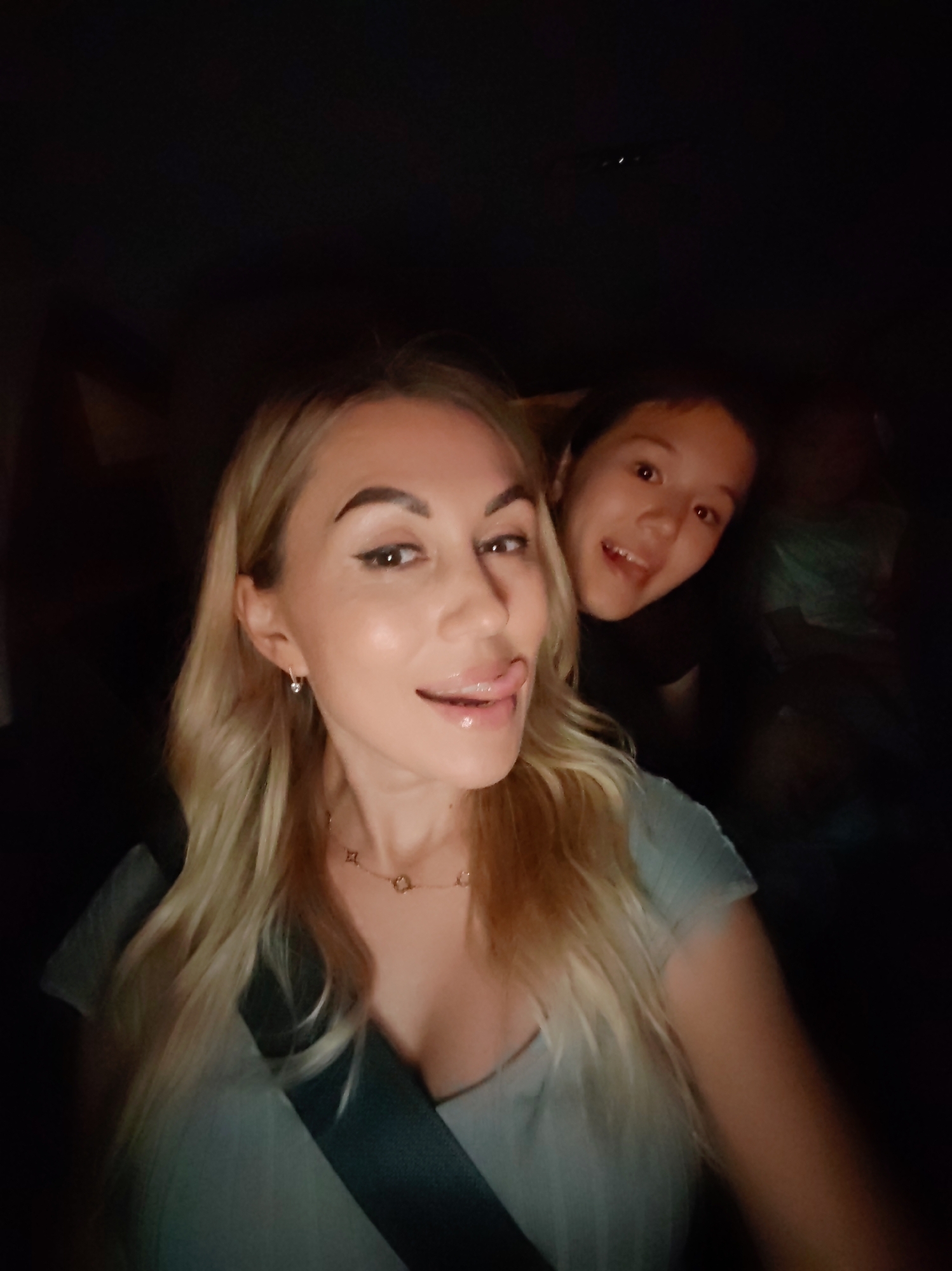 The image is a selfie taken inside a vehicle at night. Two individuals are visible. In the foreground, a blonde woman with a seatbelt across her chest is sticking her tongue out. She is wearing a light-colored top and a necklace. Behind her, a person with dark hair is looking at the camera with a wide smile. The background is dark, suggesting it is nighttime and inside a car. The location of Komsomolsk-on-Amur, Russia is not discernible from the image.