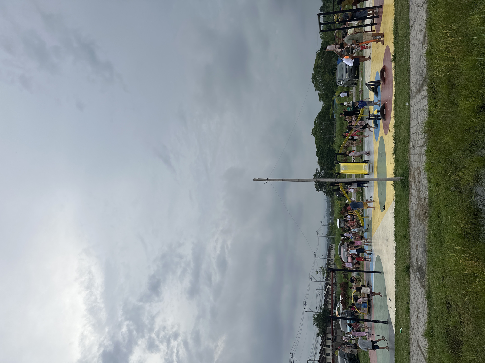 An outdoor public exercise park in Davao City, Philippines, is depicted under a heavily overcast sky. Numerous individuals, primarily adults, are engaged in physical activity across various pieces of outdoor gym equipment. The ground of the exercise area is distinctly colorful, featuring large painted circles and pathways in shades of yellow, red, blue, and light gray.
People are observed utilizing equipment such as black metal pull-up bars, leg press machines with circular components, and other fitness stations. One individual near the center wears an orange safety vest. A silver or grey car is visible near the exercise zone. A paved pathway of light grey pavers runs along the edge of the colorful ground, separating it from a strip of green grass. Beyond the grass, trees and residential structures are visible in the background, with multiple utility poles and power lines spanning the horizon. The cloudy weather suggests a mild daytime environment with diffused light. No explicit text is visible, and the scene portrays a routine public recreation and fitness setting.