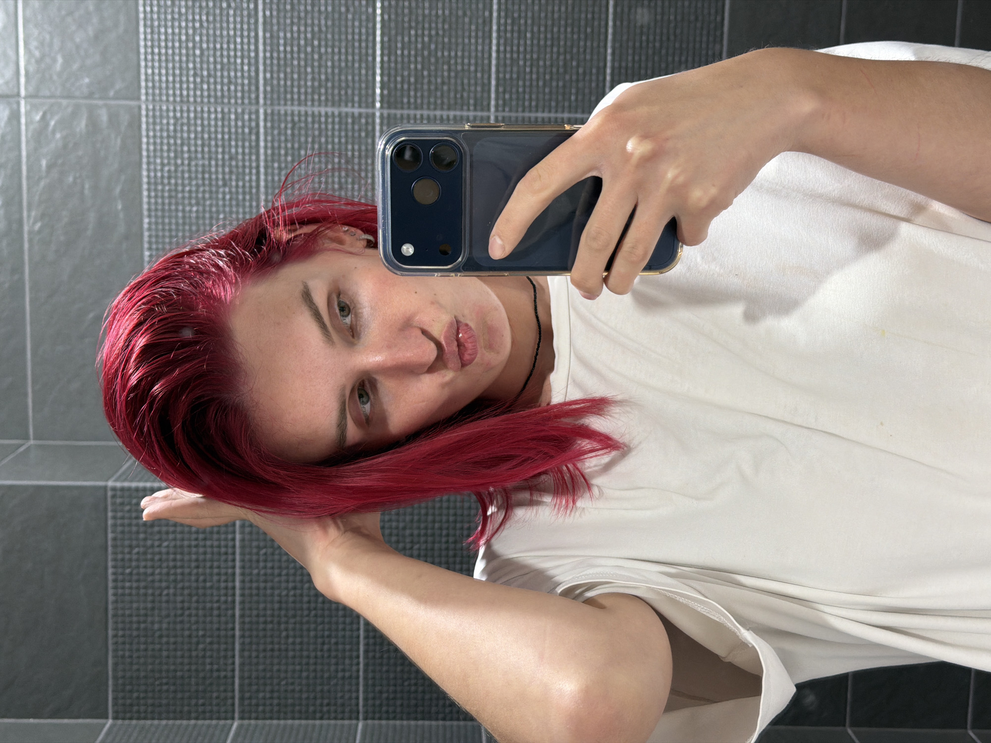 A selfie image, rotated 90 degrees clockwise, shows a young adult with vibrant red hair taking a picture in a mirror. The individual, dressed in a white t-shirt, holds a dark-colored smartphone in a clear case with gold trim in their right hand, pointing it towards their reflection. Their head is tilted, and they look towards the camera with a neutral eye expression and slightly pursed lips. A black cord or necklace is visible around their neck, and a piercing is present on their visible ear. The background is a tiled wall, typical of a bathroom or utility area, featuring two distinct grey tile patterns: an upper section with small, textured square tiles and a lower section with larger, smoother tiles that include subtle diagonal line details. No visible text is present in the image. The scene is indoors, well-lit, and the location context is Chaikovsky, Russia.