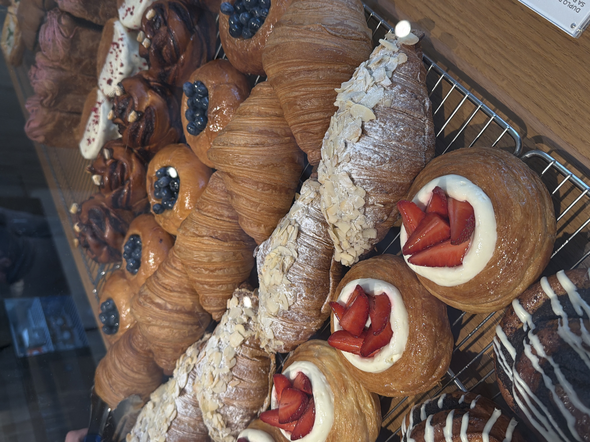 The image shows a variety of pastries arranged on a metal rack, likely displayed in a bakery or pastry shop. The pastries include croissants with almond flakes, croissants with blueberries, round pastries filled with white cream and topped with sliced strawberries, and dark pastries with white icing. The pastries are arranged closely together. The background is a wooden surface, possibly part of the display case or countertop. The reflection of a person can be seen in the glass. A partial label with the word "DUPLO" is visible. The pastries are located in Belgrade, Serbia.