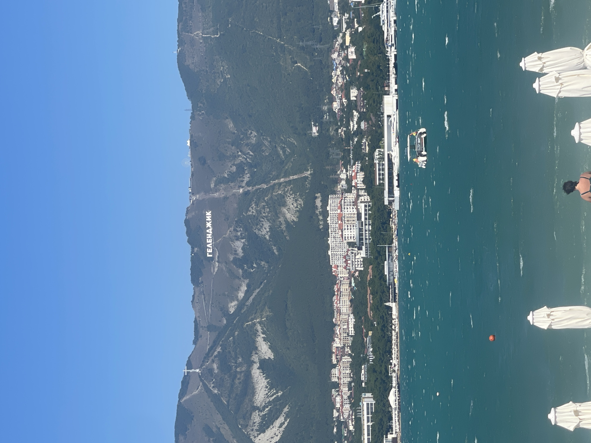 The image captures a panoramic view of Gelendzhik, Russia, with the mountains forming a dramatic backdrop to the coastal city. In the foreground, the teal waters of the sea are choppy, with whitecaps indicating a breezy day. Several closed white beach umbrellas are visible along the sandy shore, and a person wearing a dark swimsuit is standing near them. A bright orange buoy floats in the water, and a small boat with a canopy and two people on board is making its way across the sea.
The middle ground of the image showcases the vibrant cityscape of Gelendzhik. Numerous buildings with red-tiled roofs and white facades are clustered together, rising up the gentle slope towards the mountains. A promenade or waterfront area with modern structures and docked boats, including several yachts, stretches along the coastline.
Dominating the background are the imposing mountains, their slopes covered with dense green forests. High up on the mountainside, large white letters spelling out "ГЕЛЕНДЖИК" (Gelendzhik) are clearly visible, serving as a prominent landmark. A large white cross is also perched atop one of the peaks, and ski lifts or cable car tracks are visible ascending the slopes, suggesting recreational activities. The sky above is a clear, bright blue.