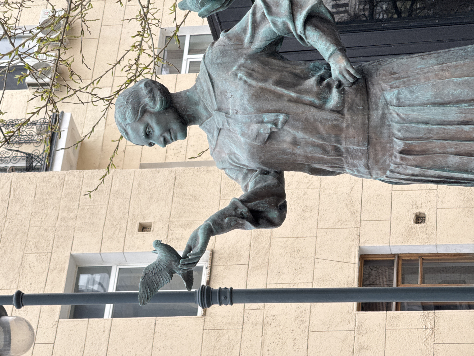 A bronze statue of a woman, facing left, is featured in front of a light-colored building. The woman is holding a bird in her outstretched left hand. The statue depicts the woman wearing a collared shirt and a long, layered skirt. Behind the statue is a building with windows and a decorative wrought-iron balcony. Bare tree branches are visible at the top left corner of the image. A metal railing and lamppost are situated in the lower part of the frame. This statue is located in Moscow, Russia.