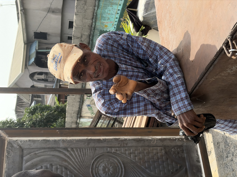 An older dark-complexioned male, wearing a light blue and dark blue plaid long-sleeved tunic and a worn light orange baseball cap with "Benson" text, is depicted leaning over a rusty, reddish-brown metallic surface in the city of Igboefon, Nigeria. His left hand, fingers slightly curled, is extended towards the viewer. His right hand grips a crumpled dark object while resting on the metallic surface. The man exhibits facial wrinkles, a mustache, goatee, and visible dental gaps. To the left, an ornate dark metal door with a sunburst and floral pattern is partially visible, adjacent to the ear and partial head of another individual. Stacks of metal rods are situated behind the man. In the midground, a concrete wall with partially visible painted text "SHARON FA" is present. The background features several multi-story concrete buildings with varied architectural details and light-colored roofs, typical of urban residential or commercial structures in Nigeria, under a bright, clear sky. Scattered green foliage and another distant green-painted wall with "SECONDAR Y" text are also visible.