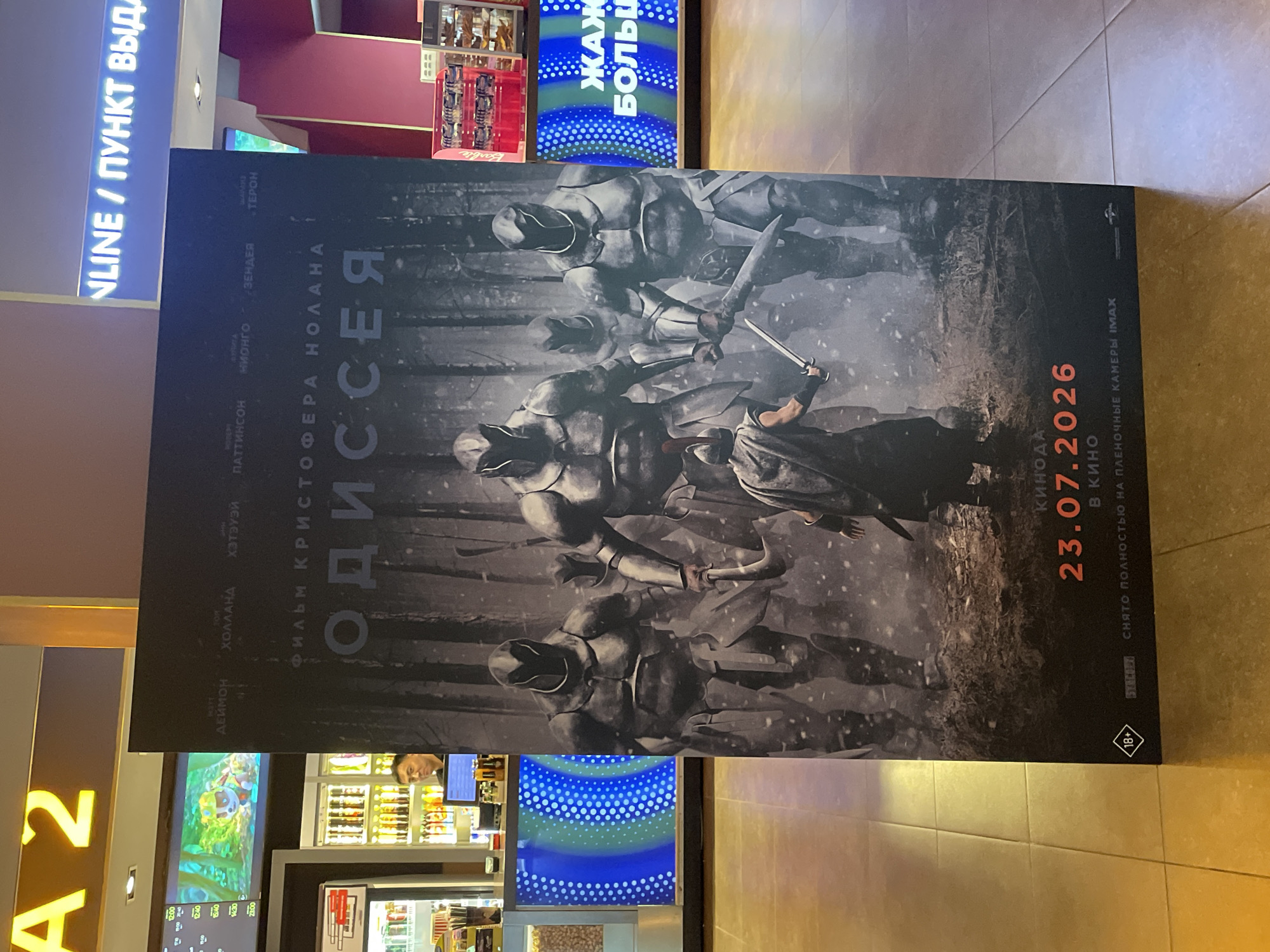 The image shows a movie poster for "Odyssey" displayed in a cinema lobby. The poster features a group of armored figures in a dark, forested setting. In the background, a concession stand is visible with a staff member behind the counter and various snacks on display. A digital display board in the concession area shows showtimes. Above the poster, a sign with yellow lettering reads "A 2," indicating the cinema or screen number. Another neon sign above the concession area reads "NLINE / ПУНКТ ВЫДА," suggesting an online service or pickup point. The floor is tiled. The poster includes the release date "23.07.2026" and mentions IMAX. The overall environment suggests a modern cinema complex. The presence of Cyrillic text on the poster and signs points to a location in a Russian-speaking country, and while not explicitly stated, it is consistent with Almaty, Kazakhstan.