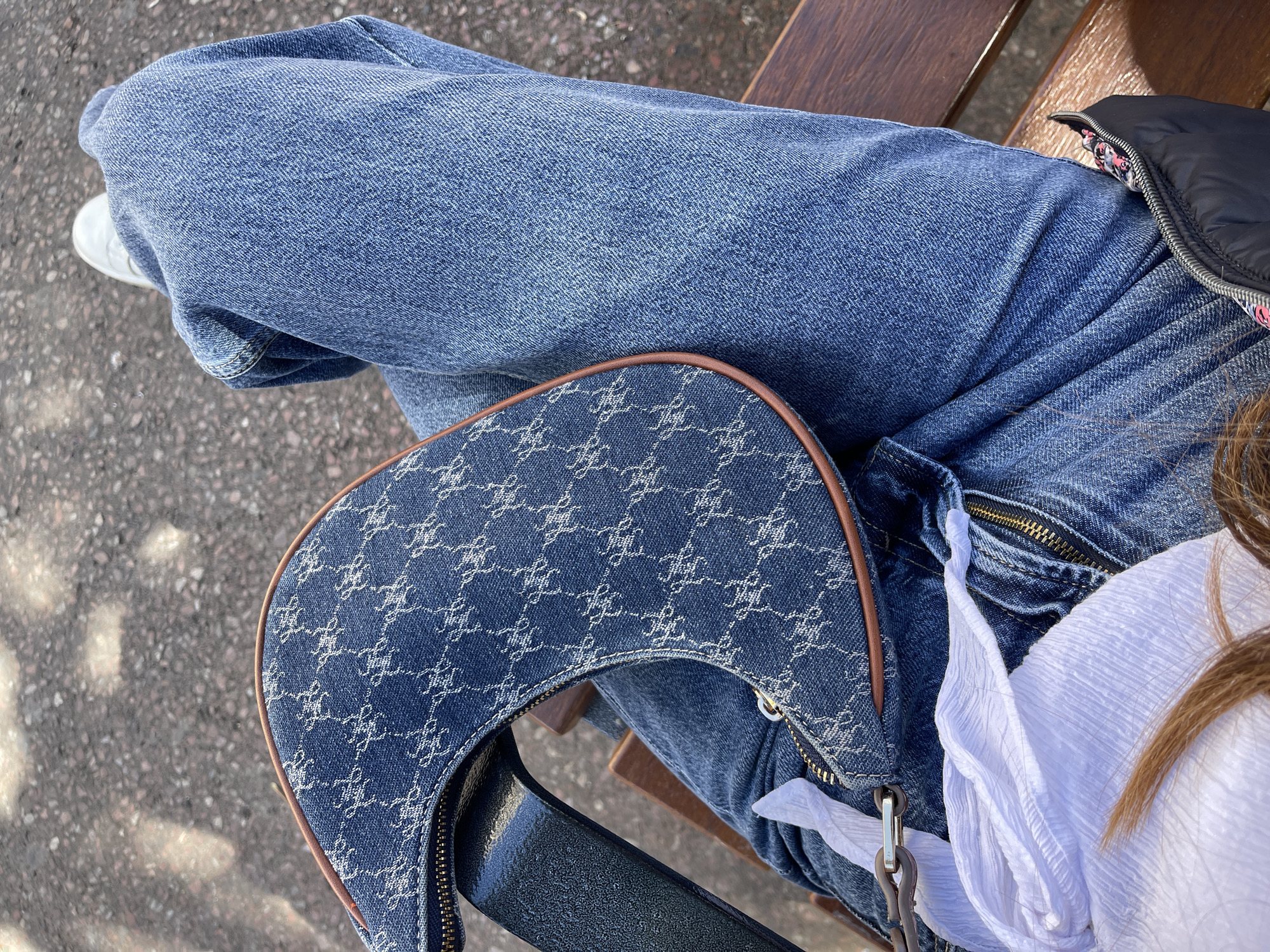 A high-angle shot shows a person seated, wearing blue jeans. A blue patterned handbag rests on their lap. A white shirt is visible on the right side of the frame. Part of a black jacket is visible in the top right corner. The person is sitting on a wooden bench. The ground below is asphalt. The location is unspecified.