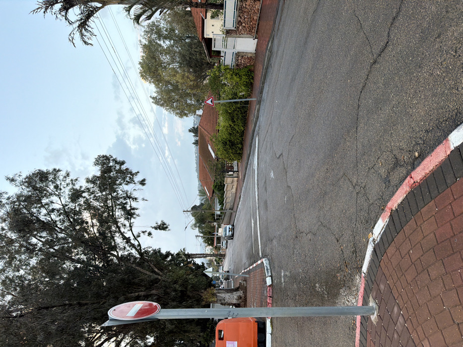 This image depicts a residential street scene in Migdal HaEmeq, Israel, under an overcast sky with patches of lighter blue. The asphalt road surface appears damp and exhibits various cracks and repairs.
In the immediate foreground on the left, a section of reddish-brown brick pavement is bordered by a red and white painted curb. A grey metallic pole rises from this pavement, supporting a circular red traffic sign with a horizontal white bar, indicating "No Entry" (R-202). Partially visible to the left of the pole is a section of an orange public container, possibly a recycling bin. A large tree with dark, dense foliage dominates the upper left quadrant.
The road curves gently to the left and extends into the midground. White painted lines traverse the road surface, consistent with the marking for a speed bump or a pedestrian crossing. Further down the street on the left, two light-colored vehicles, possibly silver or white sedans, are visible either parked or in slow motion.
On the right side of the street, a paved sidewalk runs alongside several single-story residential houses with distinctive red-tiled roofs. These properties are secured by various fencing structures, including stacked stone walls and white metal gates, interspersed with dense green bushes and trees. A red-bordered white triangular road sign, featuring a black symbol resembling a speed bump or road hump, is positioned on a pole on the right side of the road, likely warning of the traffic calming measure ahead.
Overhead, multiple electrical or telephone lines are strung across the scene. The background reveals a landscape of additional residential structures nestled among trees, stretching towards distant hills under the cloudy sky. No individuals or explicit actions or interactions are depicted within the frame.