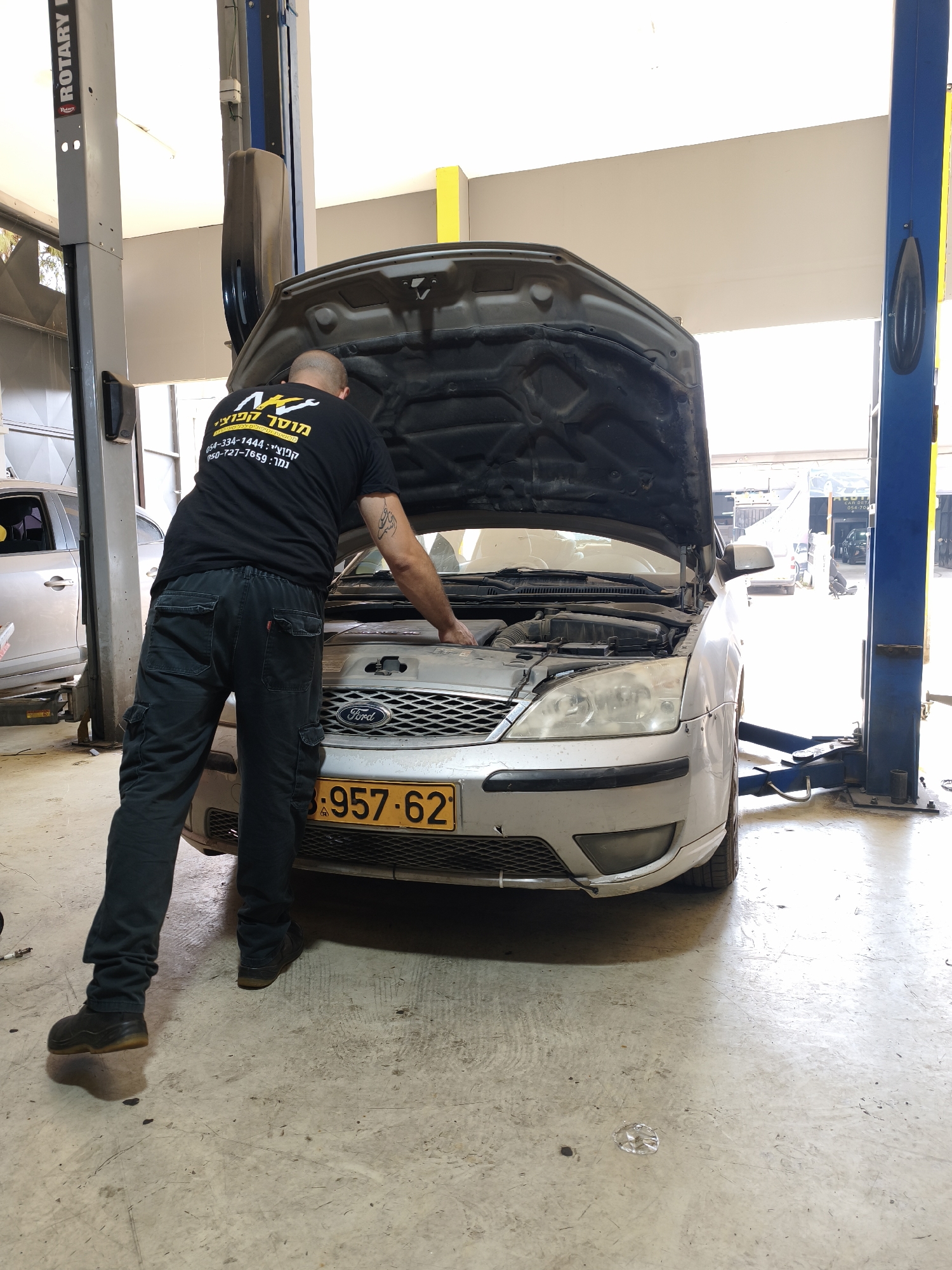 In a garage located in Kafr Yasif, Israel, a man with tattoos on his arm is working on a silver Ford car with its hood open. He is wearing a black t-shirt with a logo and text, and black cargo pants. The car is parked on a concrete floor, and a car lift is visible behind it. The garage appears to be well-lit.