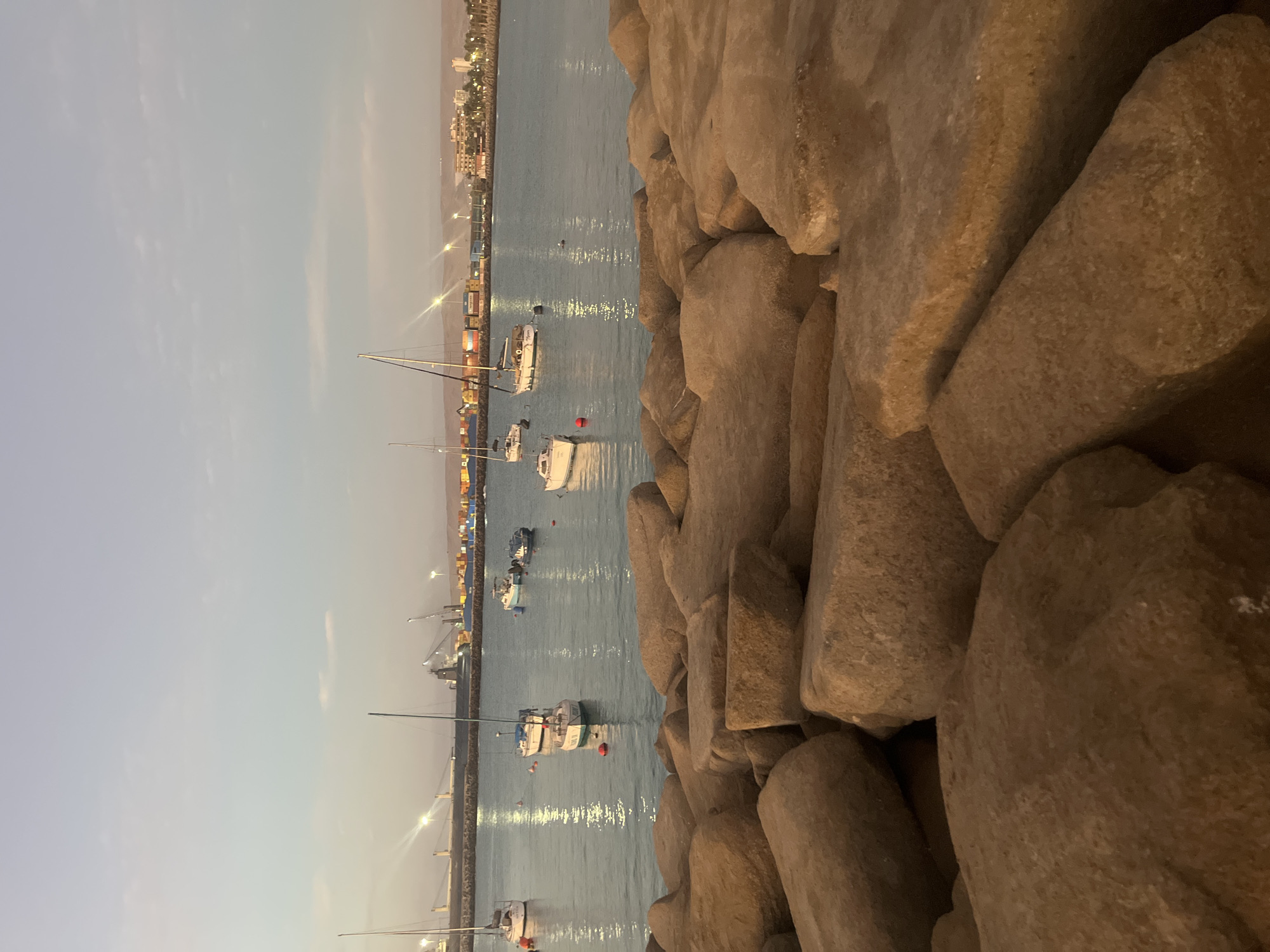 The image depicts a harbor scene in Arica, Chile, taken at dusk. The foreground consists of a rocky breakwater constructed of large, irregular boulders. Beyond the breakwater, the water is relatively calm, reflecting the soft light of the fading day and the lights from the city. Several sailboats are moored in the harbor, some with their masts clearly visible. The city of Arica stretches along the coastline in the background, with the illumination of buildings and streetlights creating a hazy glow against the darkening sky. Industrial features such as cranes and containers are also visible along the wharf.