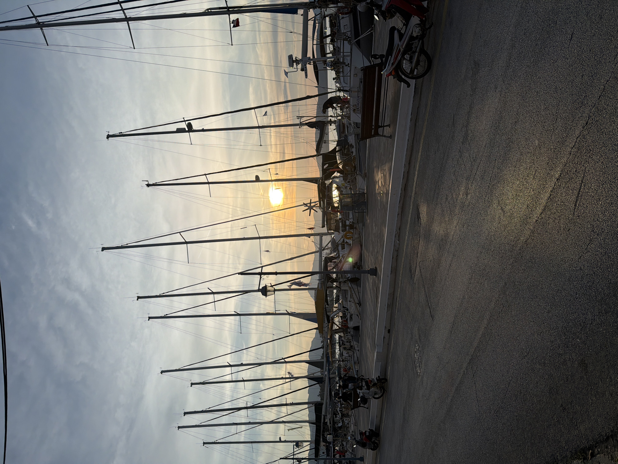 The image captures a picturesque marina scene in Poros, Greece, at either sunrise or sunset, indicated by the low-lying sun casting a golden glow across the sky. Numerous sailboats are docked along a paved waterfront, their tall masts creating prominent silhouettes against the bright sky. The sun, a brilliant disc, is positioned low on the horizon, partly obscured by the intricate rigging and masts of the boats, creating a distinct lens flare and warm illumination.
In the foreground, a dark asphalt road or pathway with a visible yellow line runs parallel to the dock. Several motorcycles or mopeds are parked along this edge, also appearing as dark silhouettes. A wooden bench is situated near one of the boats, and a lamppost is visible further down the dock.
In the background, distant mountains rise, their outlines softly defined against the lighter hues of the sky. The sky itself features a gradient of colors, transitioning from warm oranges and yellows near the sun to cooler blues and grays with scattered wispy clouds. One person is faintly discernible on a boat, but no specific activity is clearly depicted. On the hull of one of the larger sailboats, the name "PATAGON" is visible. The scene evokes a tranquil maritime atmosphere, characterized by strong backlighting and dramatic contrasts.