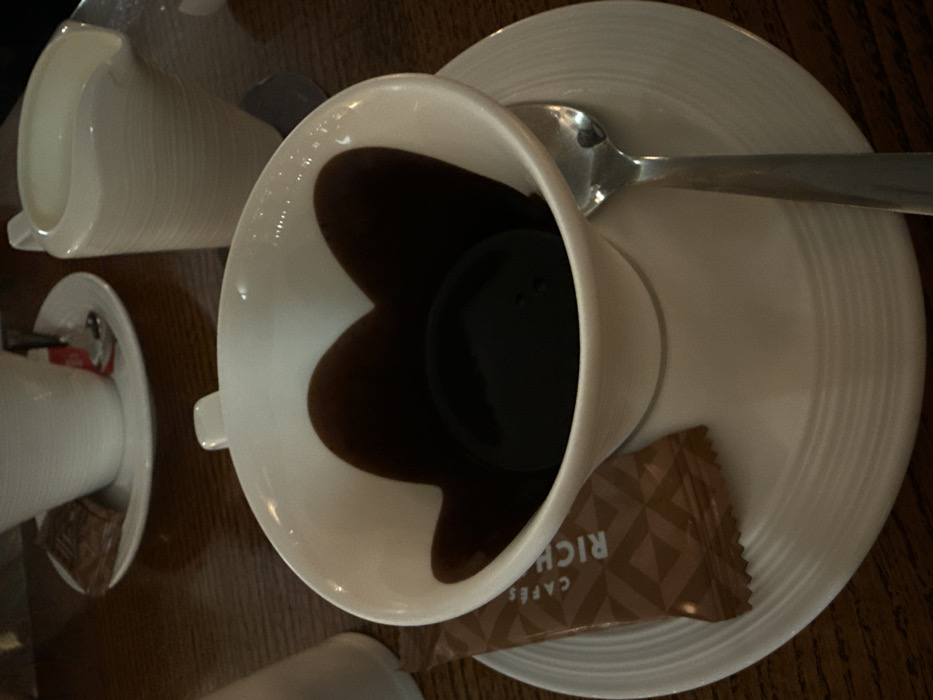 A white teacup sits on a matching saucer, filled with dark coffee that appears heart-shaped. A silver spoon rests on the saucer's edge. A small packet of "Rich Cafe" sugar is placed near the teacup. A similar, empty teacup and saucer are visible to the left of the main set. The scene is set on a wooden surface.