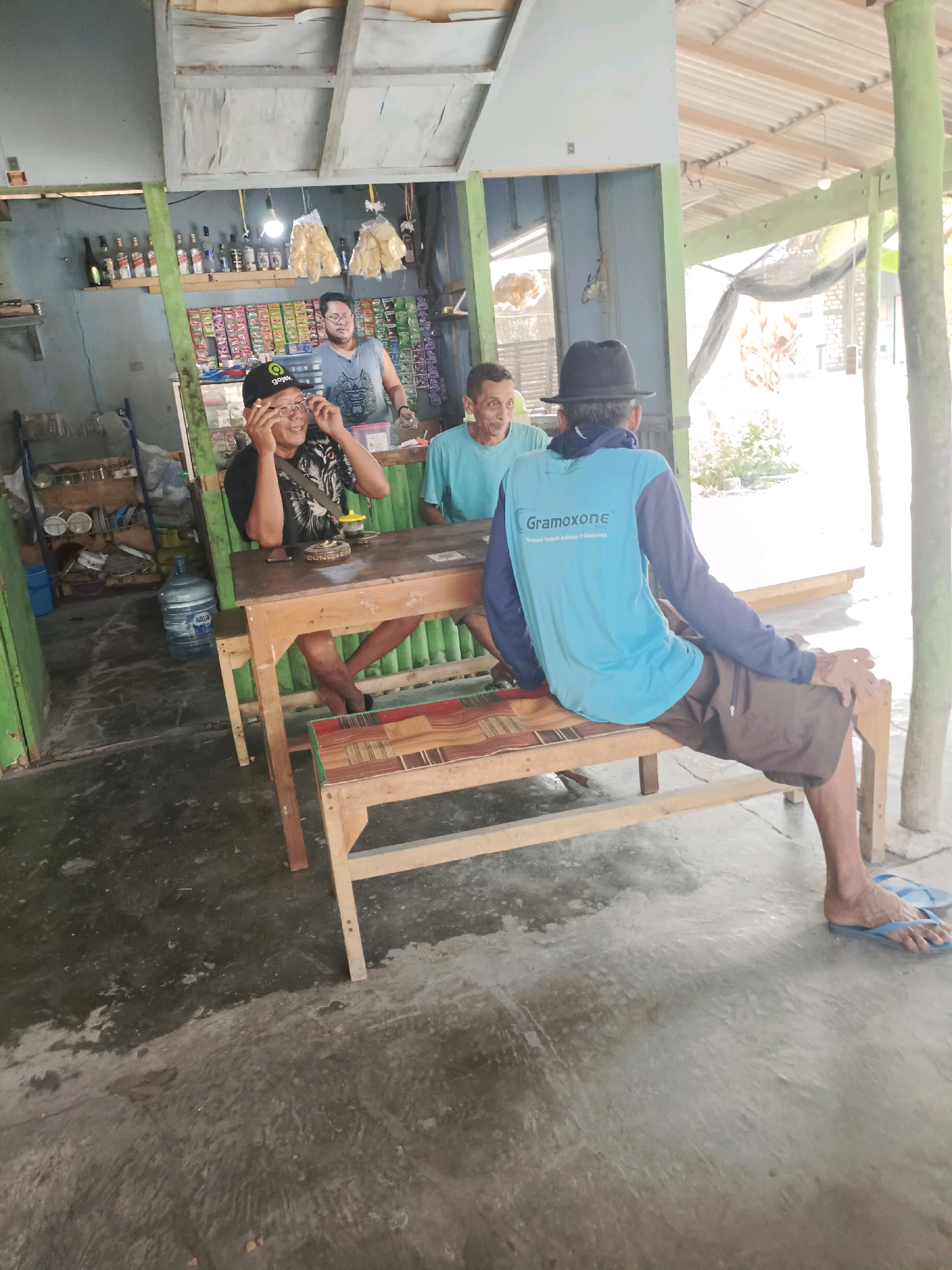 In Rancang Kencono, Indonesia, four men are gathered at a wooden table in what appears to be an outdoor or semi-outdoor establishment with a corrugated metal roof and a concrete floor. Two of the men are seated at the table, facing away from the camera. One man, wearing a black hat and a blue long-sleeved shirt with the text "Gramoxone" on the back, is seated on a bench. He is wearing brown shorts and blue flip-flops. The other man seated at the table, facing the camera, wears glasses and a black t-shirt with a tiger graphic. He is gesturing with his hands near his face, possibly adjusting his glasses.
Behind the table, two other men are visible. One man, wearing a light blue t-shirt, is seated at the table and looking towards the man in glasses. Another man stands further back, wearing a sleeveless blue shirt and glasses, and looking towards the front. The background reveals shelves stocked with bottles and packaged goods, suggesting a small shop or warung. A large water dispenser is also present to the left of the table. The lighting is natural, with some overhead illumination.