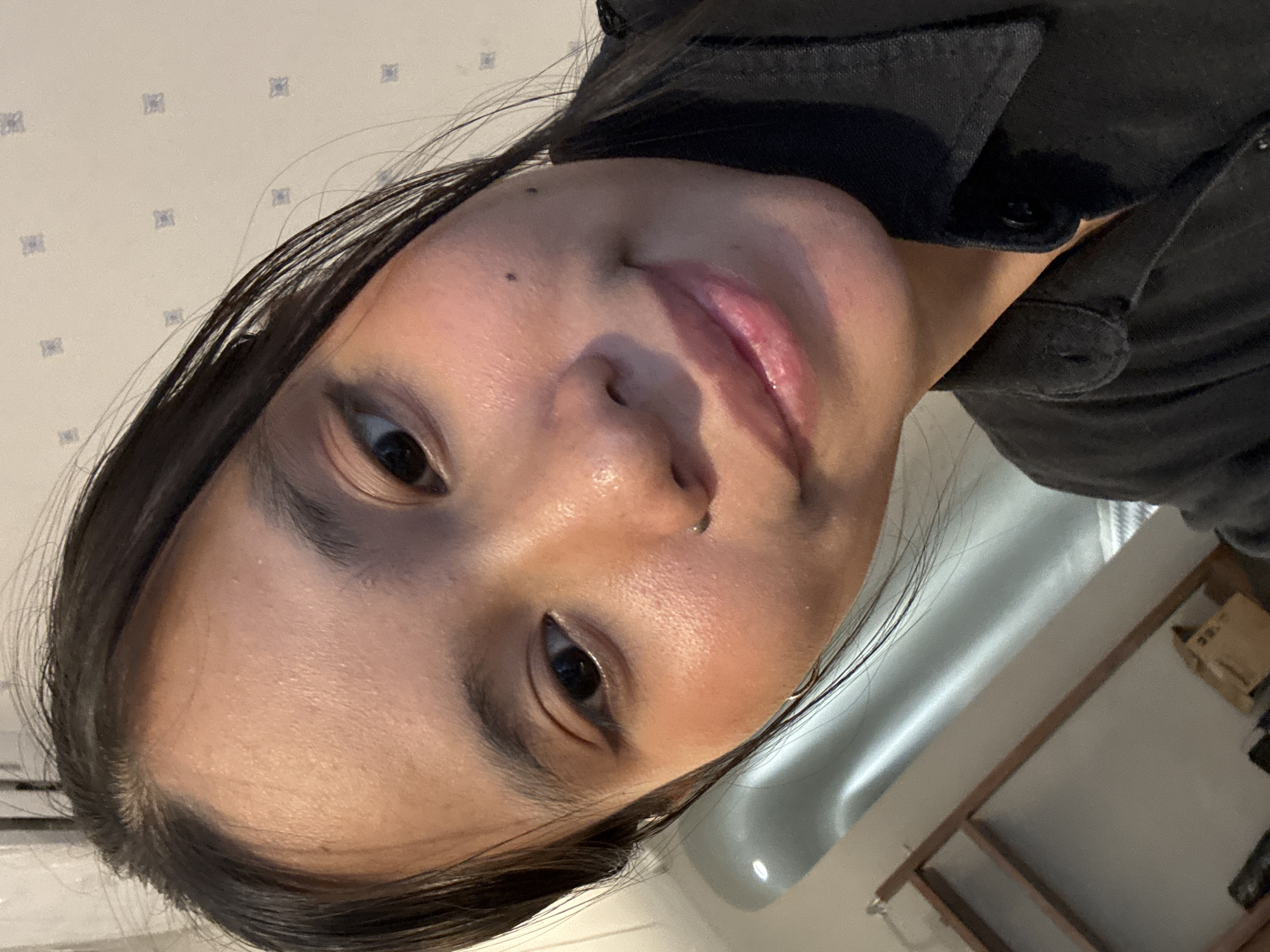 The media file is a close-up, rotated portrait of a young person with dark hair, likely taken indoors. The subject is looking towards the camera, with their face tilted. They have dark eyes, visible eyebrows, and visible pores and skin texture. There is a small dark mole on their forehead and a nose ring. Their lips are slightly parted, and they appear to be wearing light makeup. They are wearing a dark-colored garment, possibly a shirt or jacket, with visible buttons.
The background includes a textured wall with a subtle repeating pattern, possibly wallpaper. To the right of the subject, there is a light-colored, curved surface that could be part of a sink or bathtub. Further in the background, there is a wooden shelf unit with some items on it, including a cardboard box. The lighting suggests an indoor environment, and the overall atmosphere is casual and personal, typical of a selfie. There is no visible text in the image. The location context provided is South Jakarta, Indonesia, which offers no specific visual cues within the image itself. The image does not provide information about the time of day or weather.